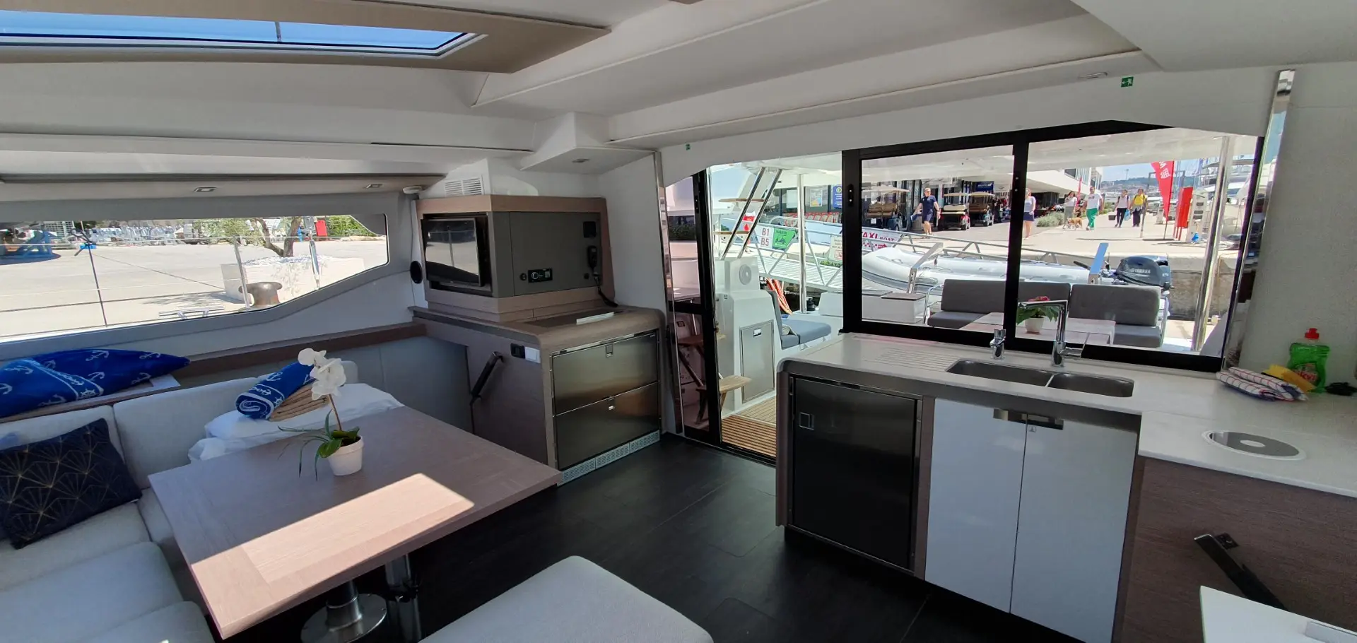 Interior of a modern Fountaine Pajot Elba 45 yacht with a dining table, cushioned seating, kitchen appliances, and a view out the window to a dockside area with boats and people outside.