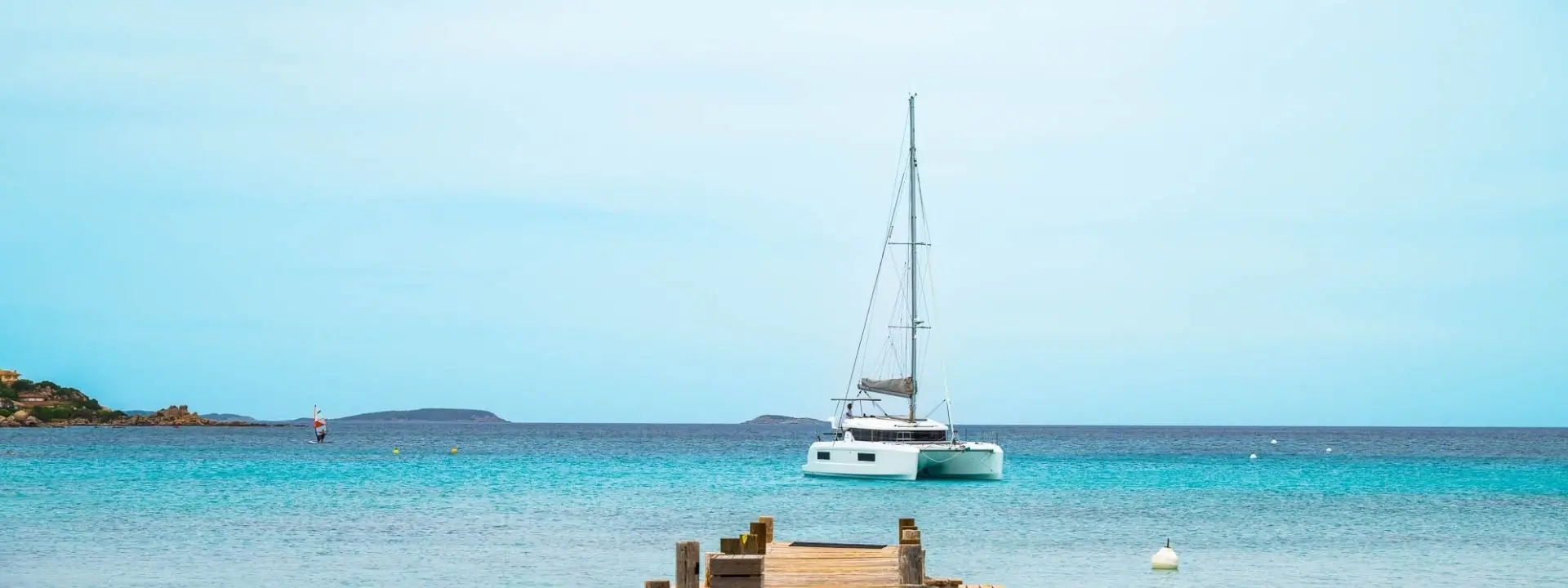 A white Lagoon 38 sailboat is anchored near a wooden pier in calm, turquoise water under a clear blue sky. Small islands and buoys are visible in the distance.