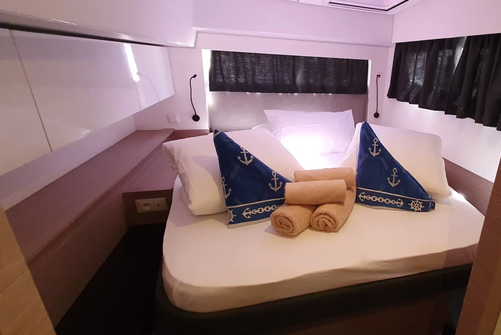 A neatly made bed in a small, cozy room aboard the Fountaine Pajot Elba 45 features two blue anchor-themed pillows and three rolled tan towels. The space has white walls, dark curtains, and soft lighting for a relaxing ambiance.