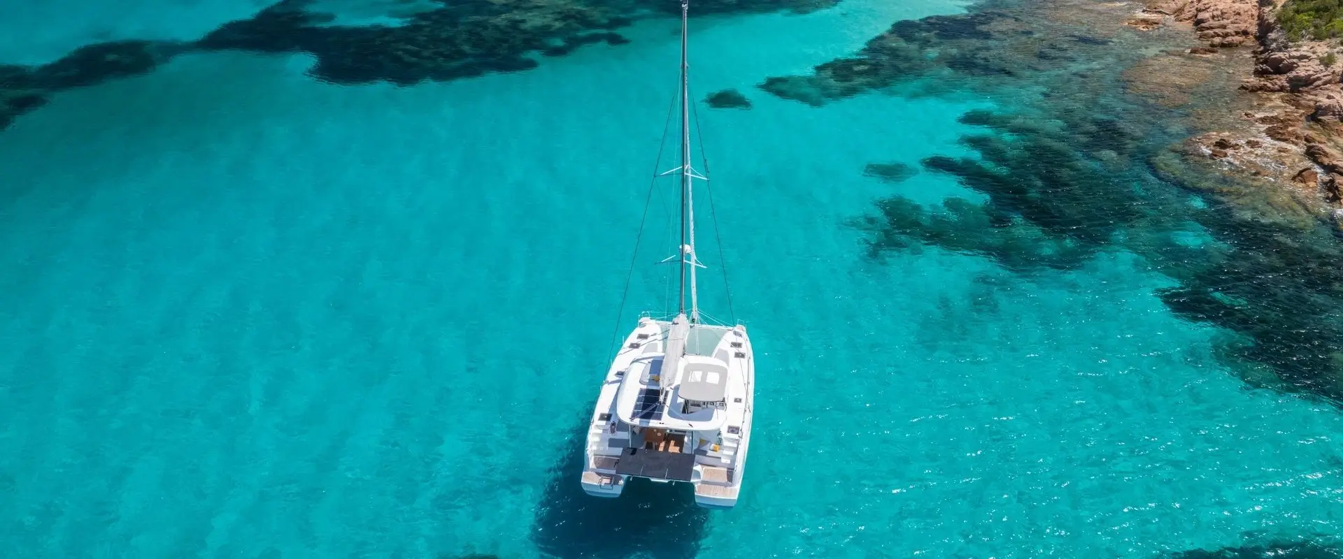 A white Lagoon 38 catamaran floats on clear turquoise water near a rocky shoreline, with dark patches of underwater vegetation visible below the surface.