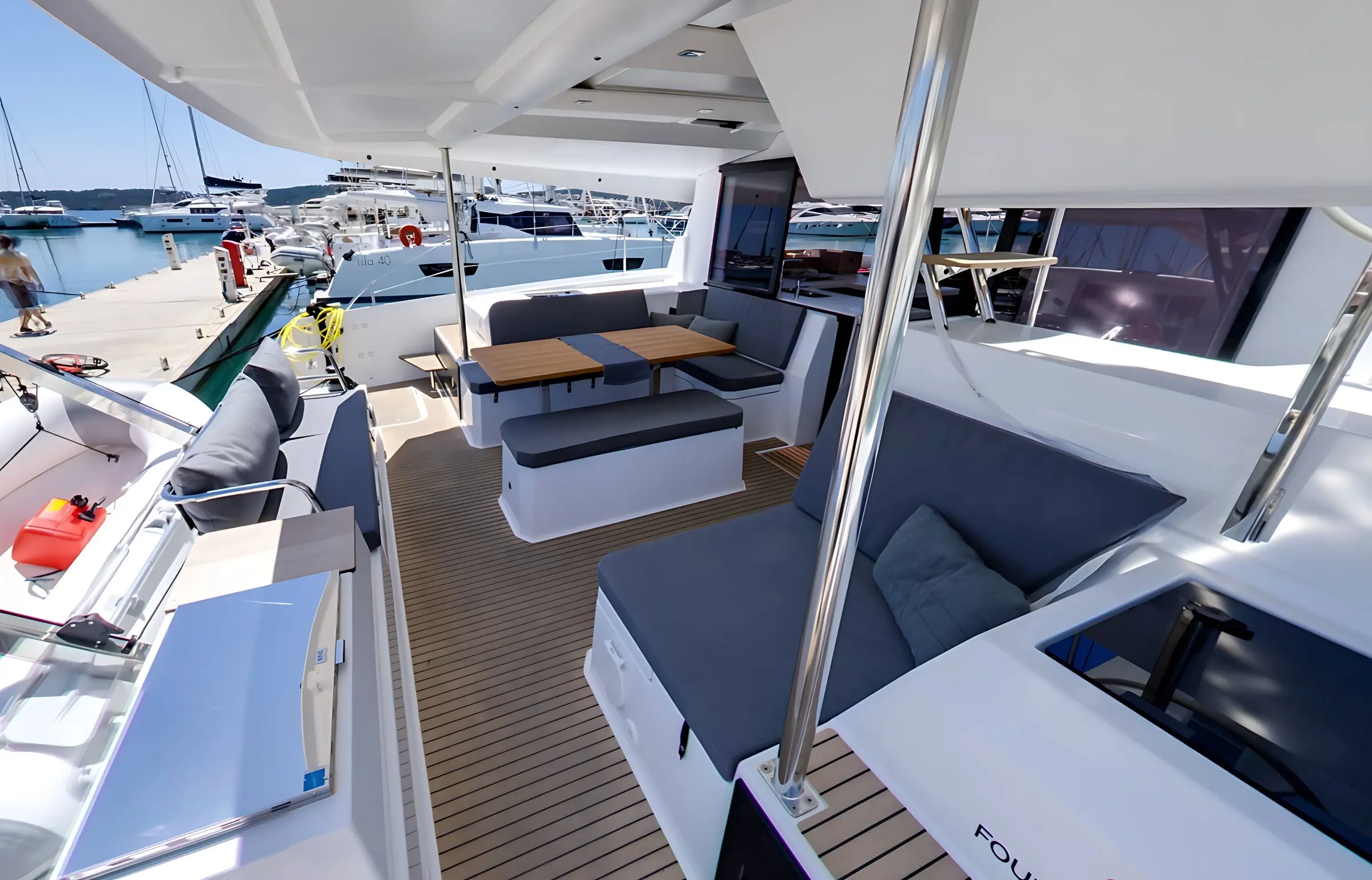Spacious Fountaine Pajot Astrea 42 yacht deck with cushioned seating, wooden tables, and metal railings; docked at a marina with several other boats and a clear blue sky in the background.