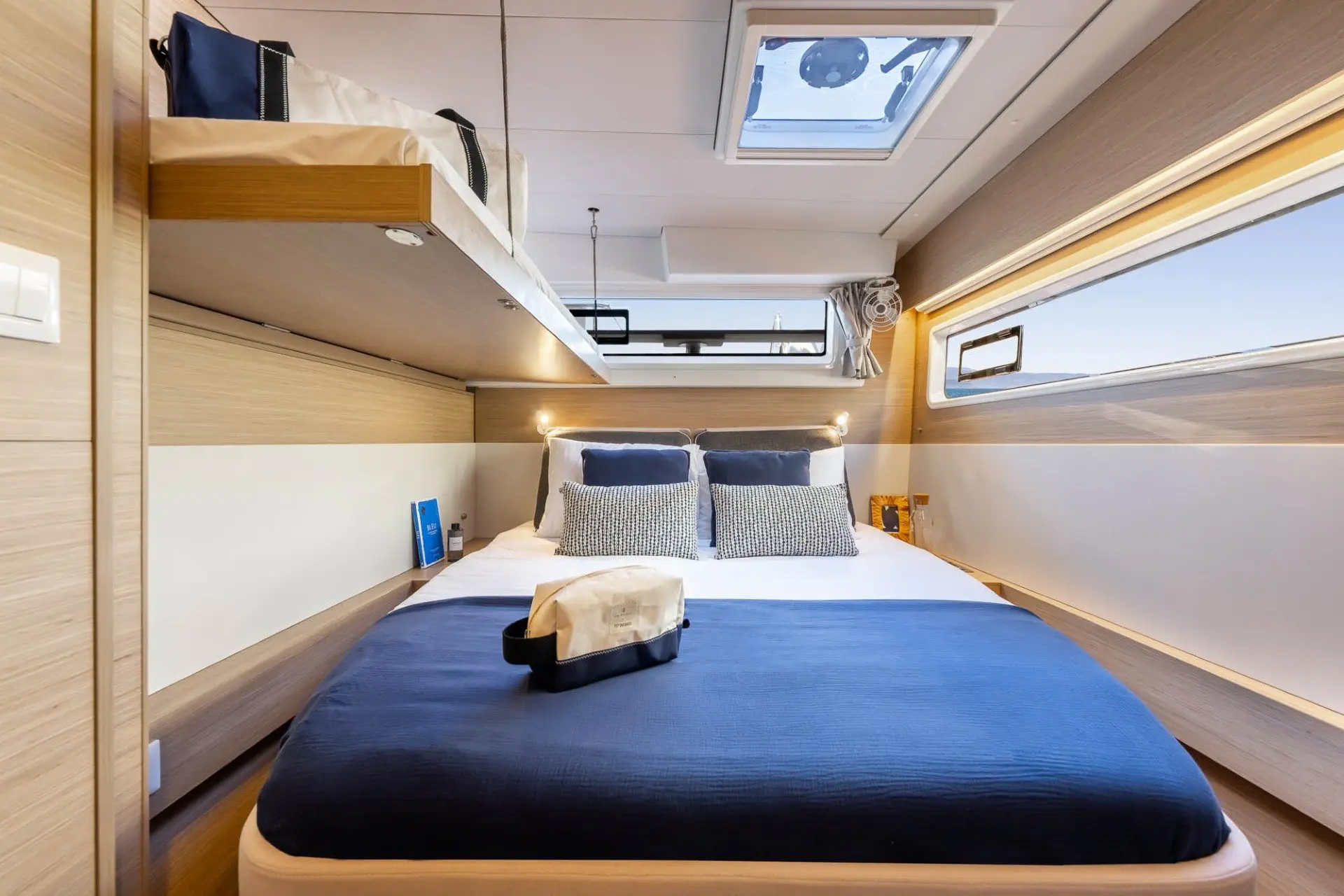 Bright, modern cabin with a double bed dressed in white and navy bedding, overhead bunk, reading lights, windows, and neatly arranged toiletries on the bed, featuring light wood paneling and a skylight above.