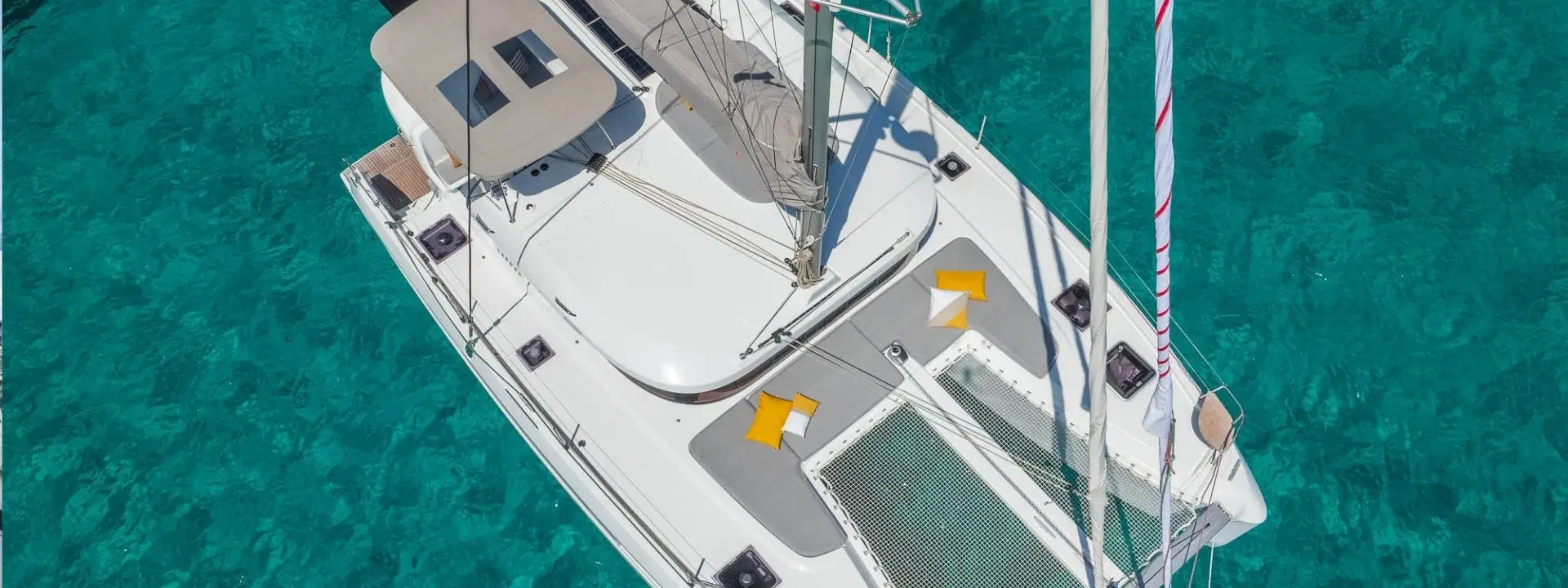 Aerial view of a Lagoon 38 sailboat with tan cushions and yellow pillows, floating on clear turquoise water. The boat’s deck and netting are visible, and sunlight reflects off the water below.