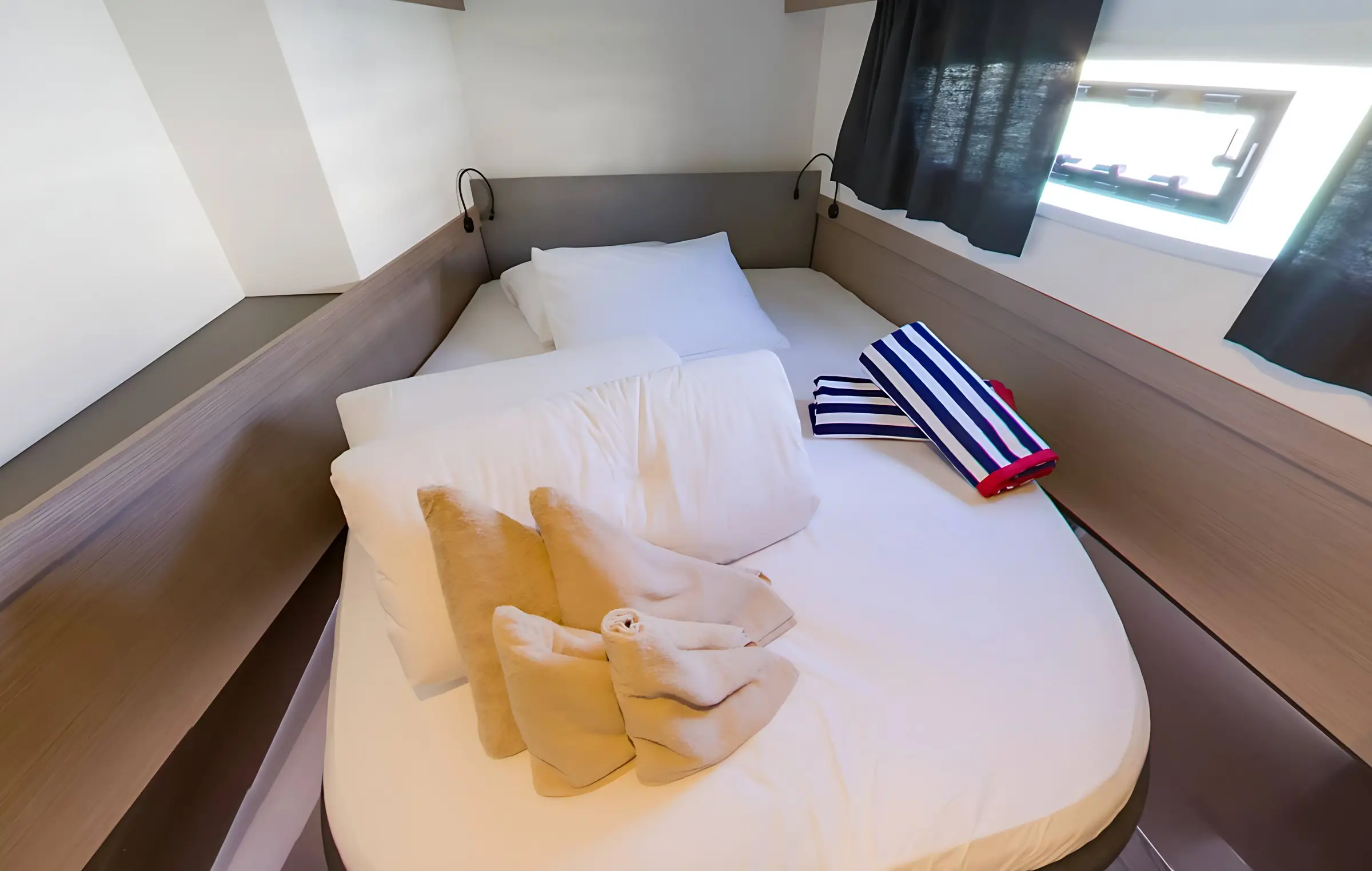 A neatly made bed in a small, modern room aboard the Fountaine Pajot Astrea 42, with white and beige bedding, two towel swans, and two rolled striped towels beside a window with dark curtains.
