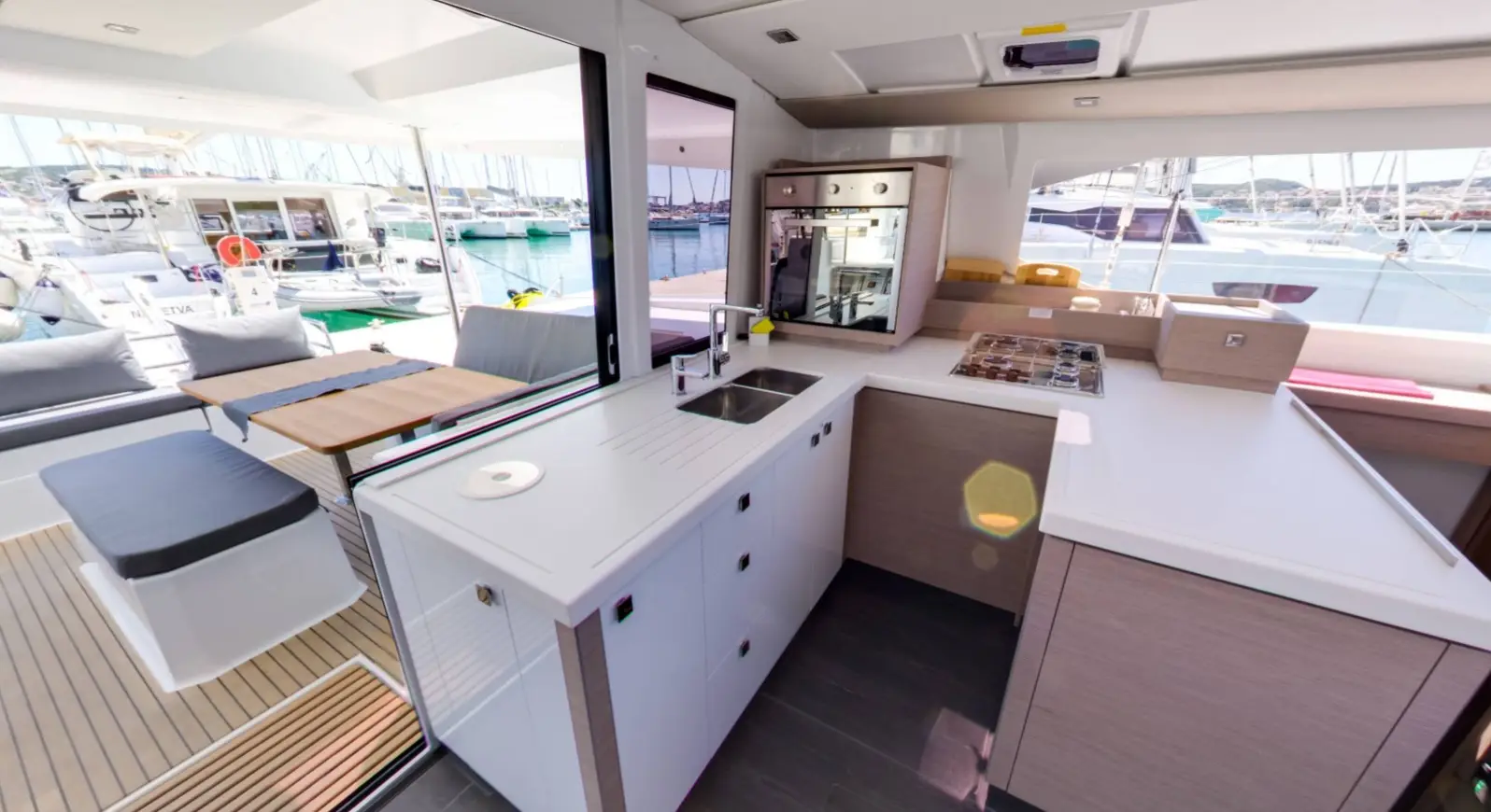 Modern yacht interior of a Fountaine Pajot Astrea 42 featuring a compact kitchen with white cabinets, sink, stove, and oven. Large windows overlook a marina with boats. Adjacent seating has a wooden table and cushioned bench. Bright, airy atmosphere.