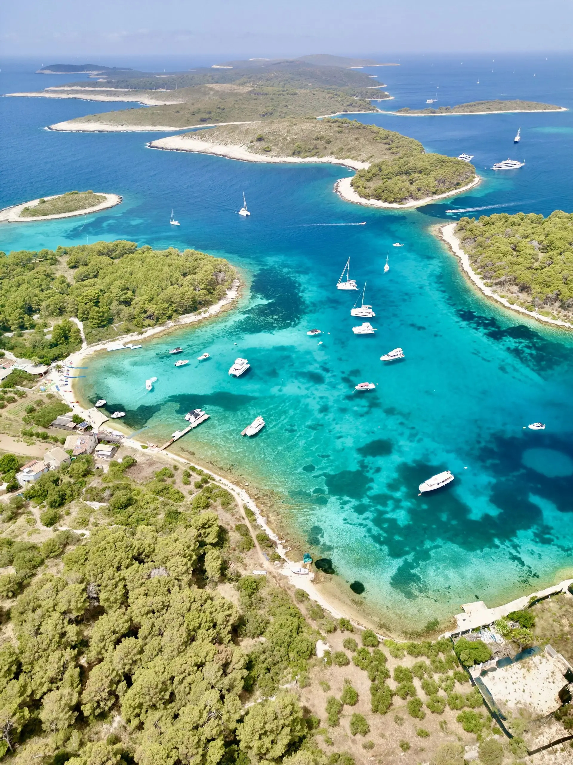 Aerial view of a turquoise bay with Lagoon 46 catamarans, sailboats, and motorboats, surrounded by lush green islands and clear blue sea under a partly cloudy sky. Sandy and rocky beaches line the shorelines.
