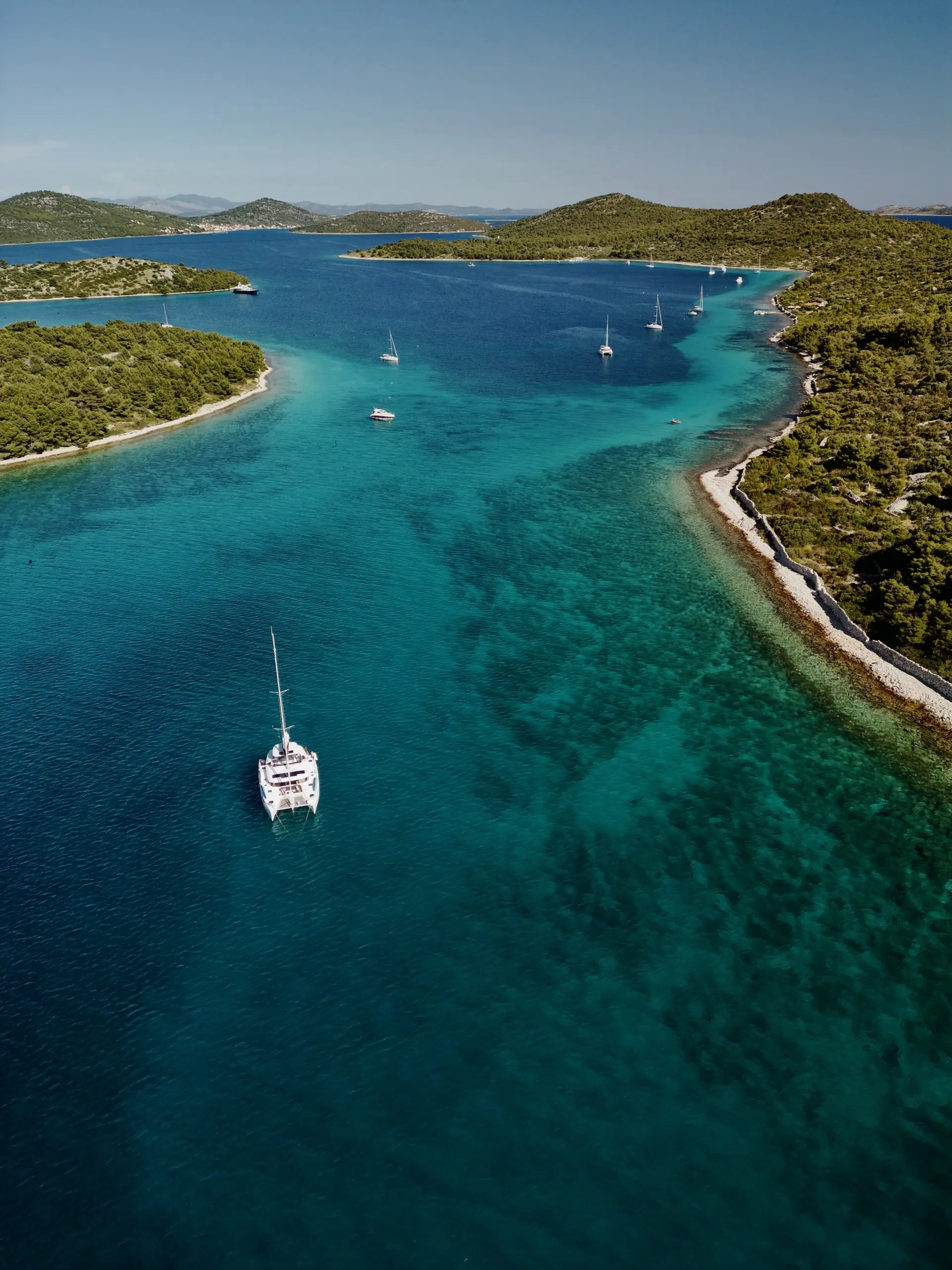 Aerial view of a Lagoon 46 catamaran sailing in clear turquoise water, surrounded by green hills and several other sailboats in the distance under a blue sky.