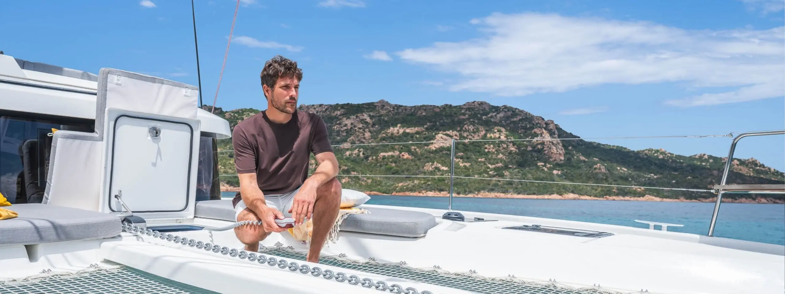 A man sits relaxed on the deck of a Lagoon 38 catamaran, surrounded by blue water and rocky, tree-covered hills under a partly cloudy sky.
