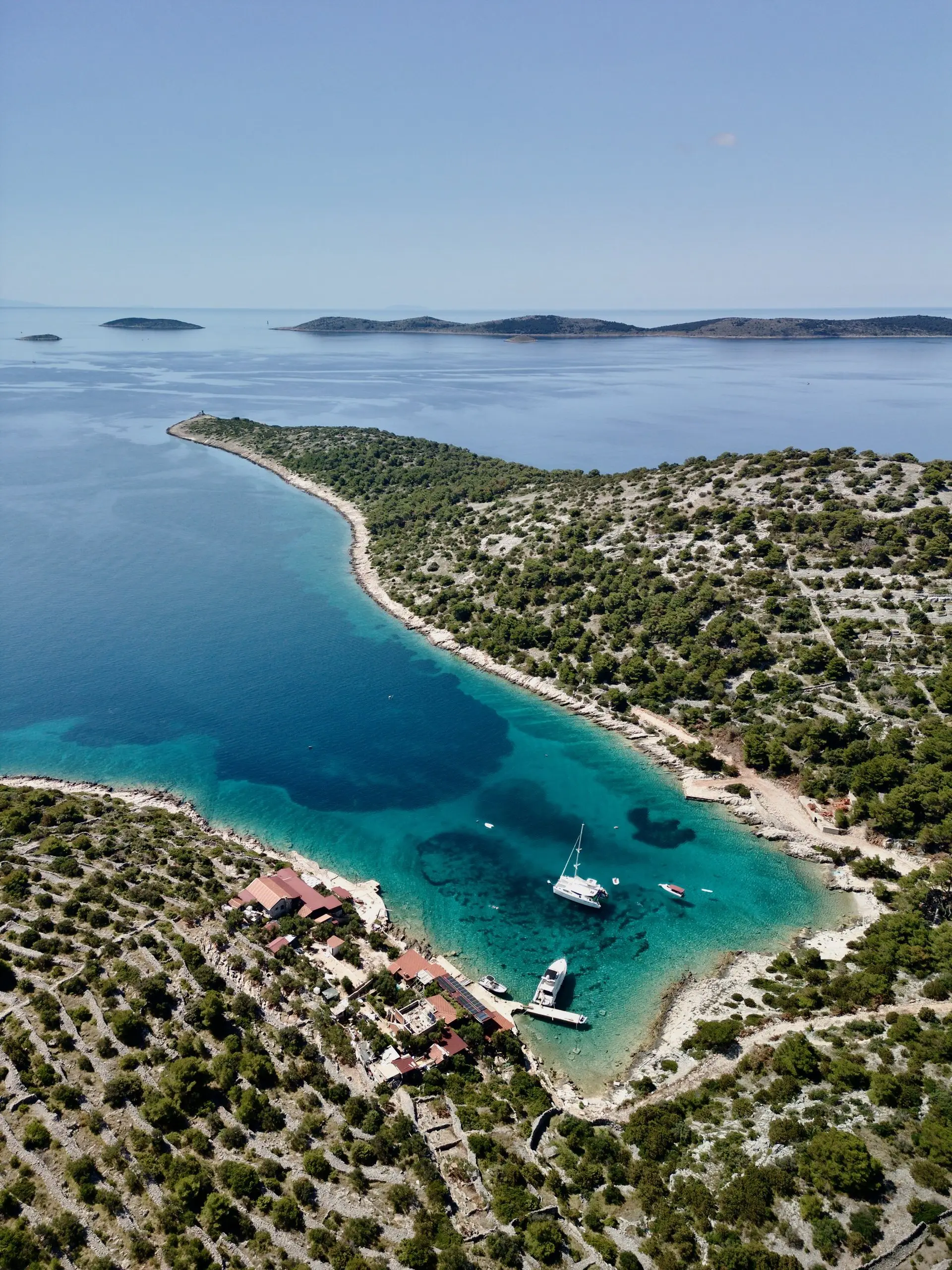 Aerial view of a narrow turquoise bay surrounded by green, hilly coastline. Several boats, including a Lagoon 46, are anchored near a small cluster of buildings at the water’s edge. Distant islands and calm blue sea stretch into the horizon.