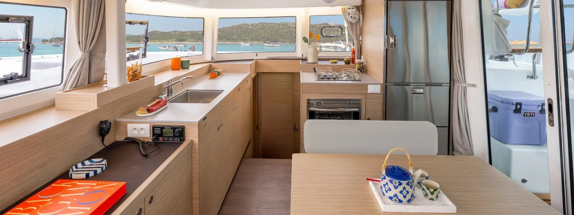 Modern Lagoon 38 yacht kitchen and dining area with wood cabinets, stainless steel appliances, and large windows overlooking a scenic bay with boats and hills. A table is set with a teapot and cups.