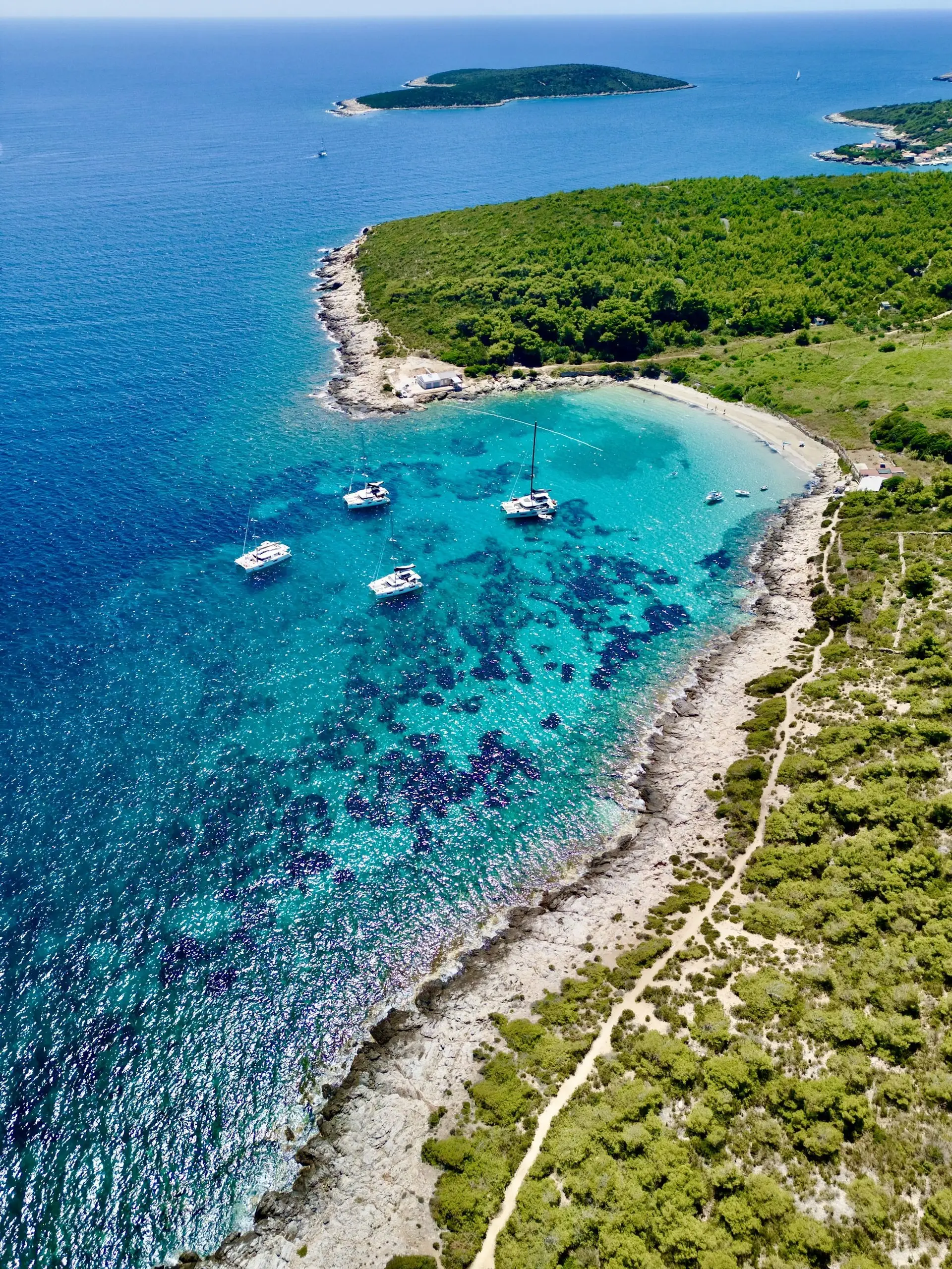 Aerial view of a turquoise bay with four sailboats, including a Lagoon 46, anchored near a rocky, forested coastline. A narrow path winds through lush vegetation along the shore, with distant islands under a clear sky.
