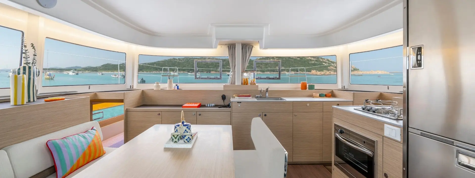 Modern Lagoon 38 yacht kitchen and dining area with light wood cabinets, stainless steel appliances, and large windows offering views of the sea and distant hills. Bright cushions and table decor add a colorful accent.