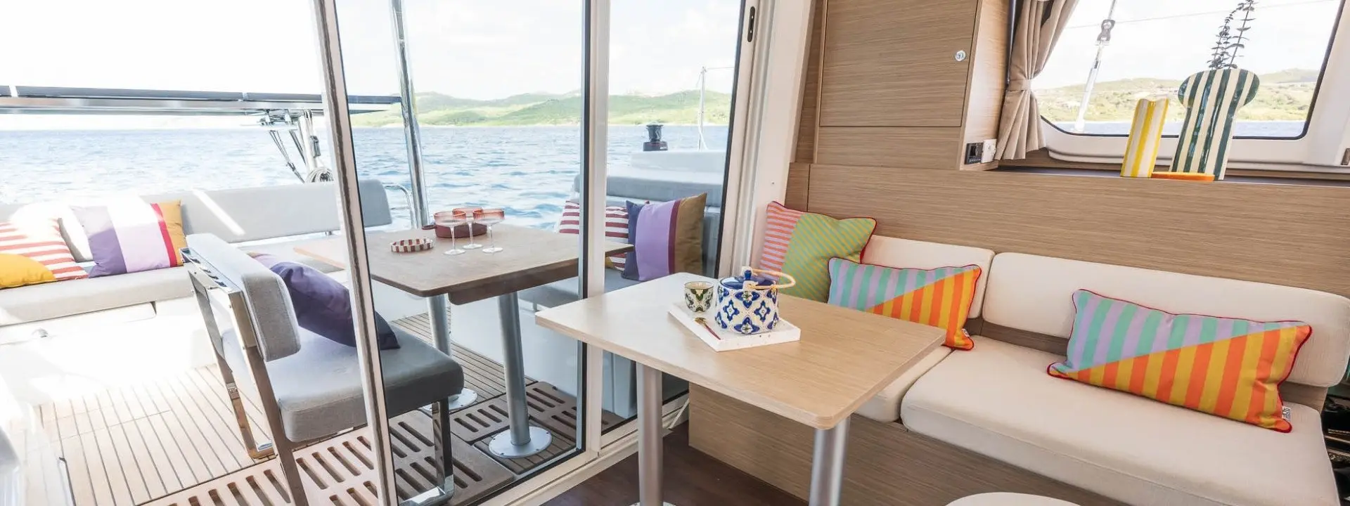 Modern Lagoon 38 yacht interior with a cozy seating area, colorful striped pillows, small wooden tables, and decorative items. Large windows provide a view of blue water and distant green hills.