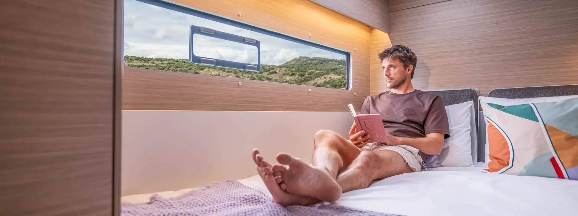 A man sits on a bed in a modern camper van, reading a book. He is barefoot and relaxed, enjoying the tranquil vibe reminiscent of lounging aboard a Lagoon 38, with a large window framing scenic hills and greenery outside.