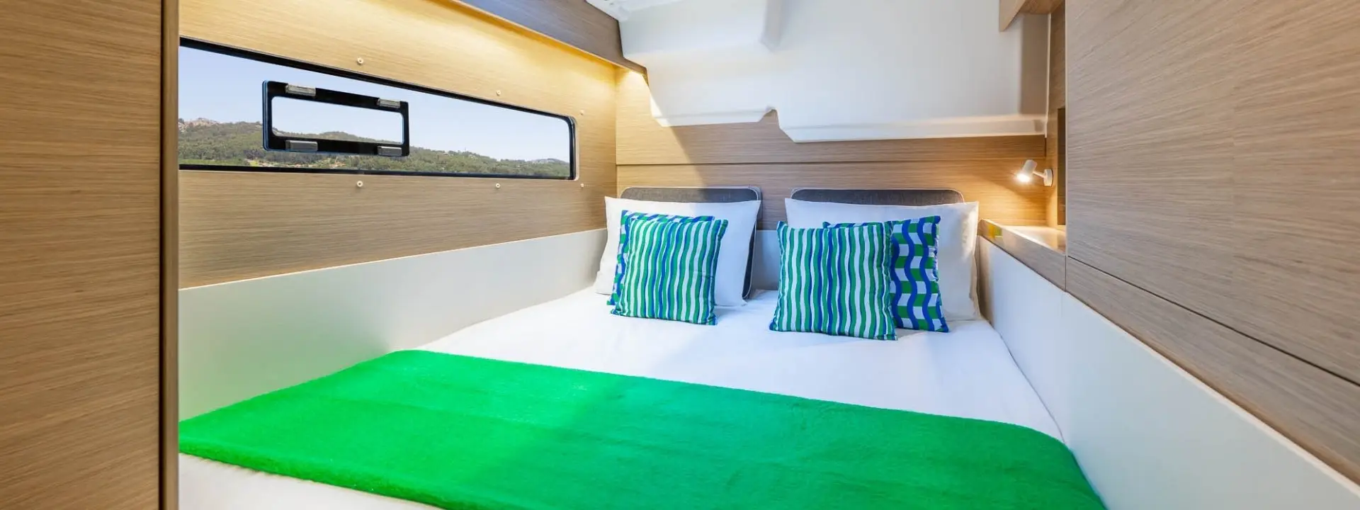 A cozy Lagoon 38 bedroom with light wood paneling, a large bed with white linens, green and blue accent pillows, a green throw, and a window offering a scenic outdoor view.