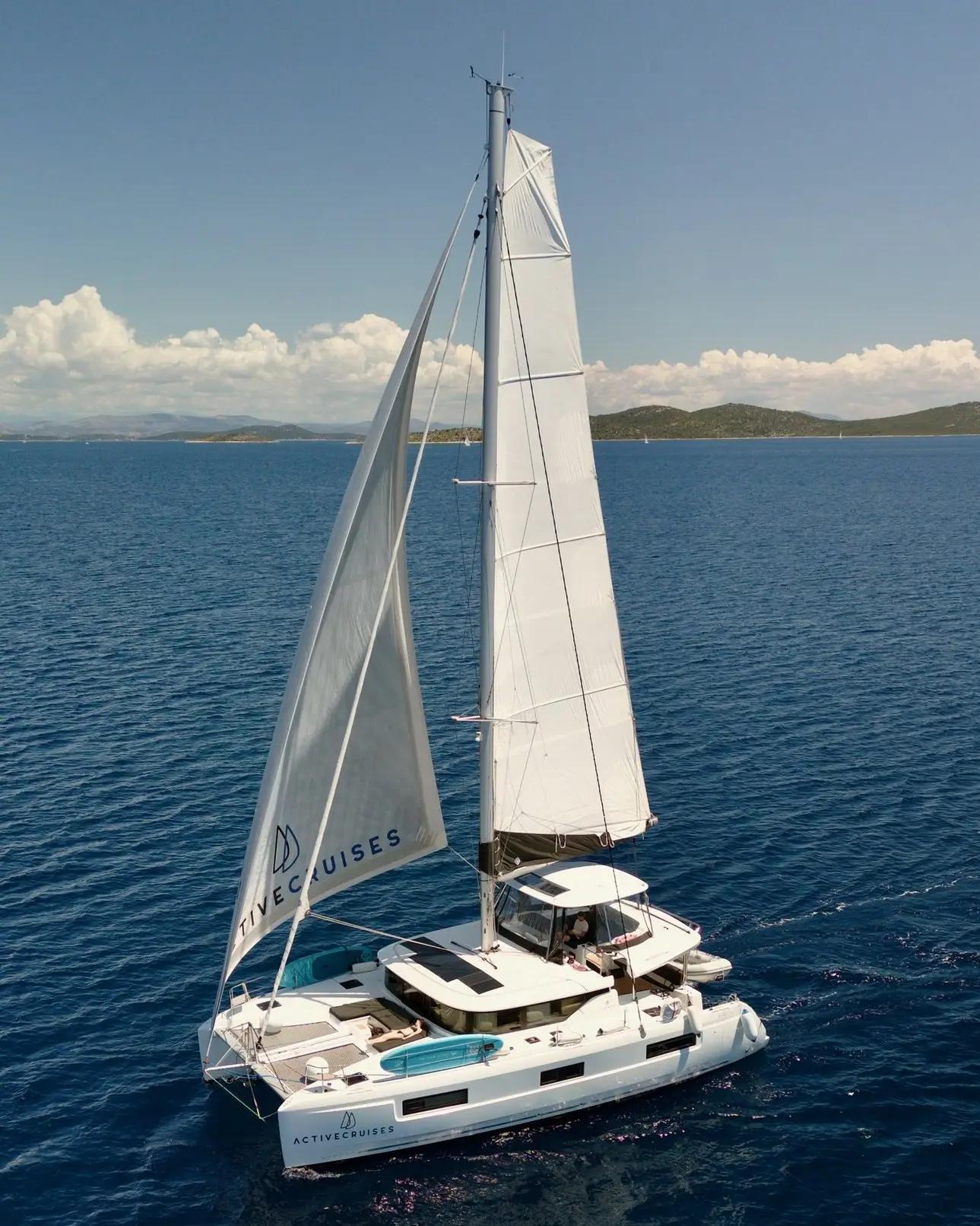 A white Lagoon 46 catamaran sailboat with raised sails glides on calm, deep blue sea under a clear sky. Hills and clouds are visible in the distance on the horizon.