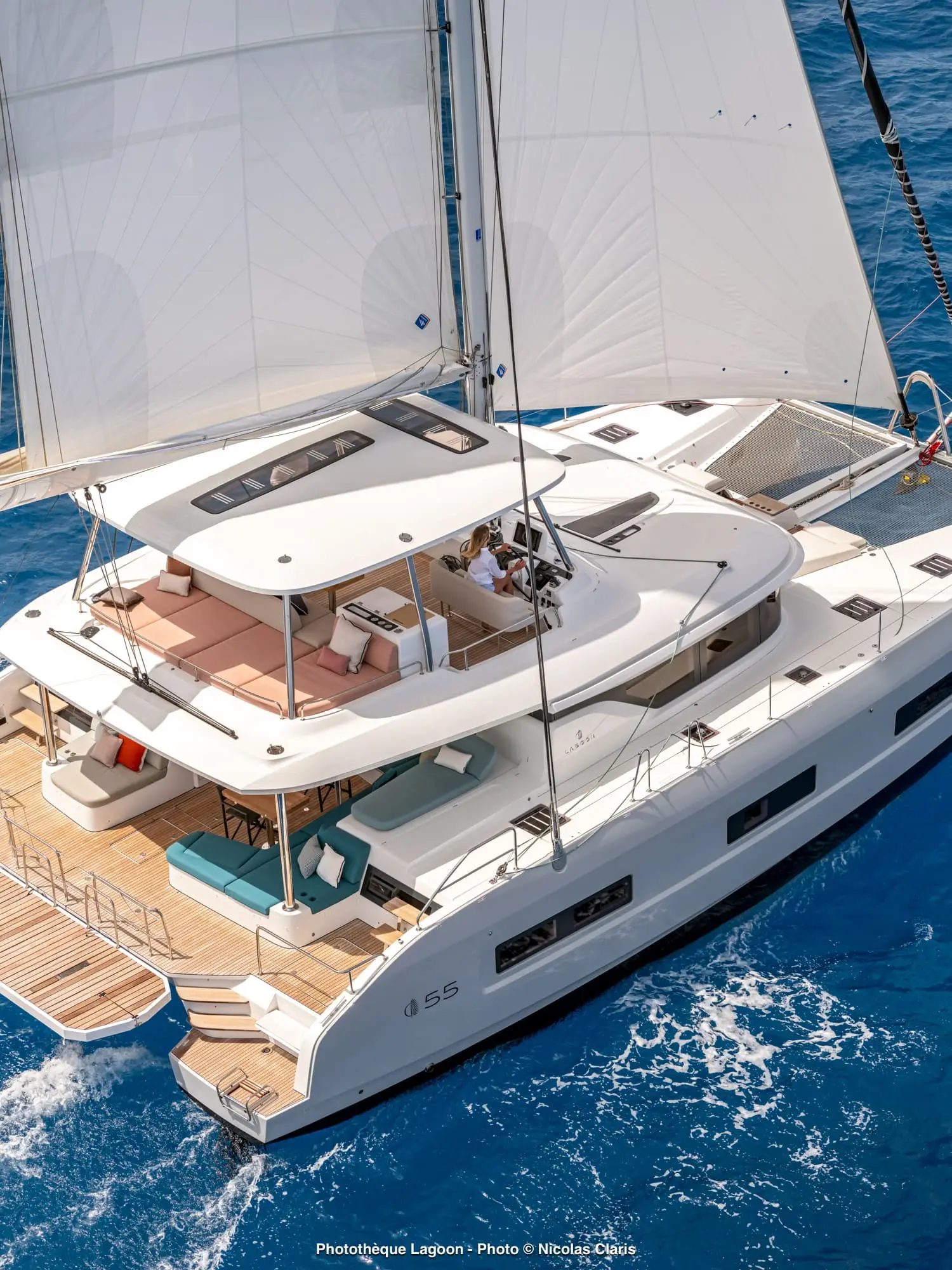 A white Lagoon 55 catamaran yacht with large sails glides on bright blue water. People relax on the upper deck, which features cushioned seating and a shaded area. The lower deck has lounge chairs and open space.