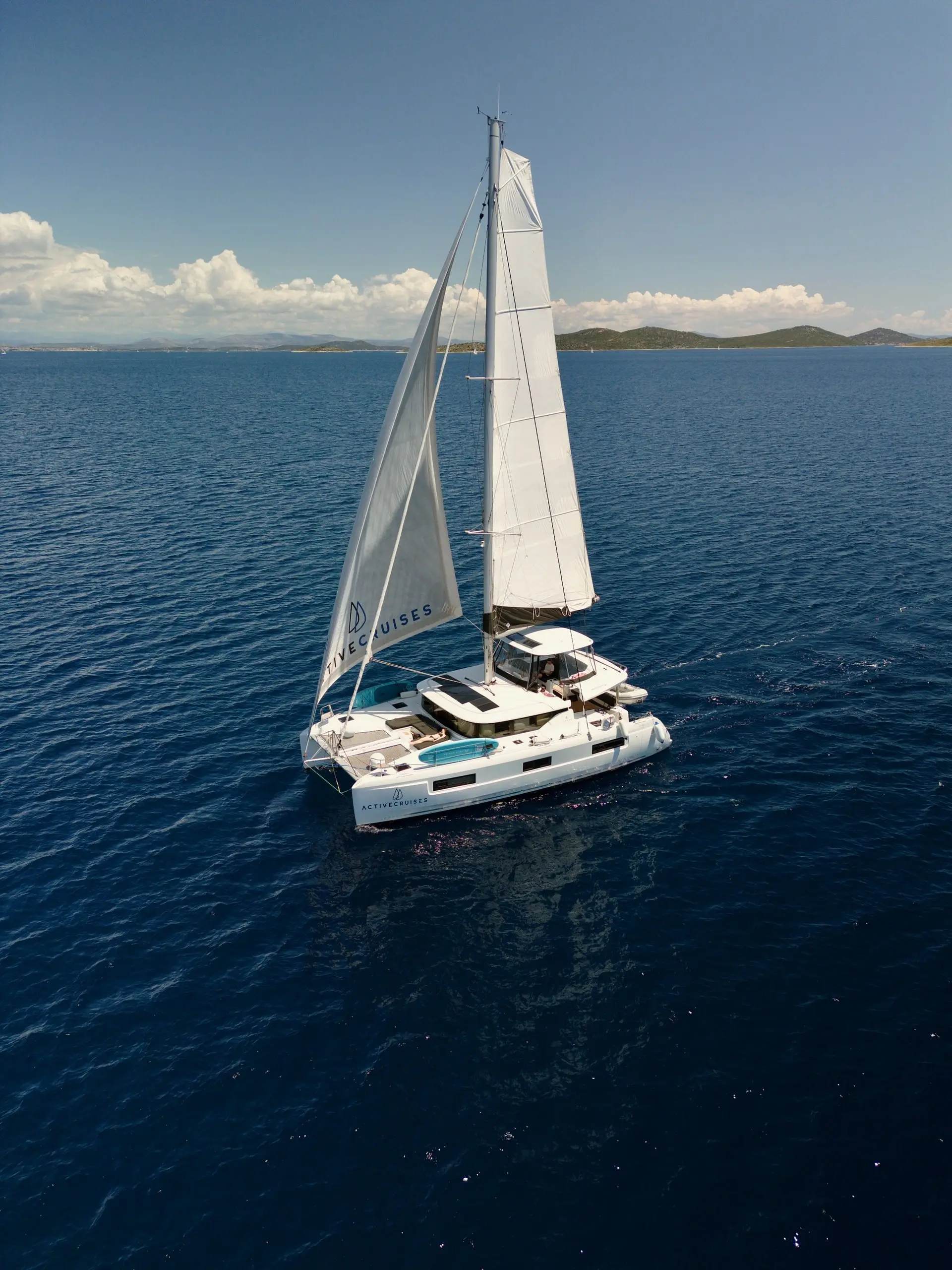 A white Lagoon 46 catamaran sailboat with raised sails glides on calm, deep blue sea under a clear sky. Hills and clouds are visible in the distance on the horizon.