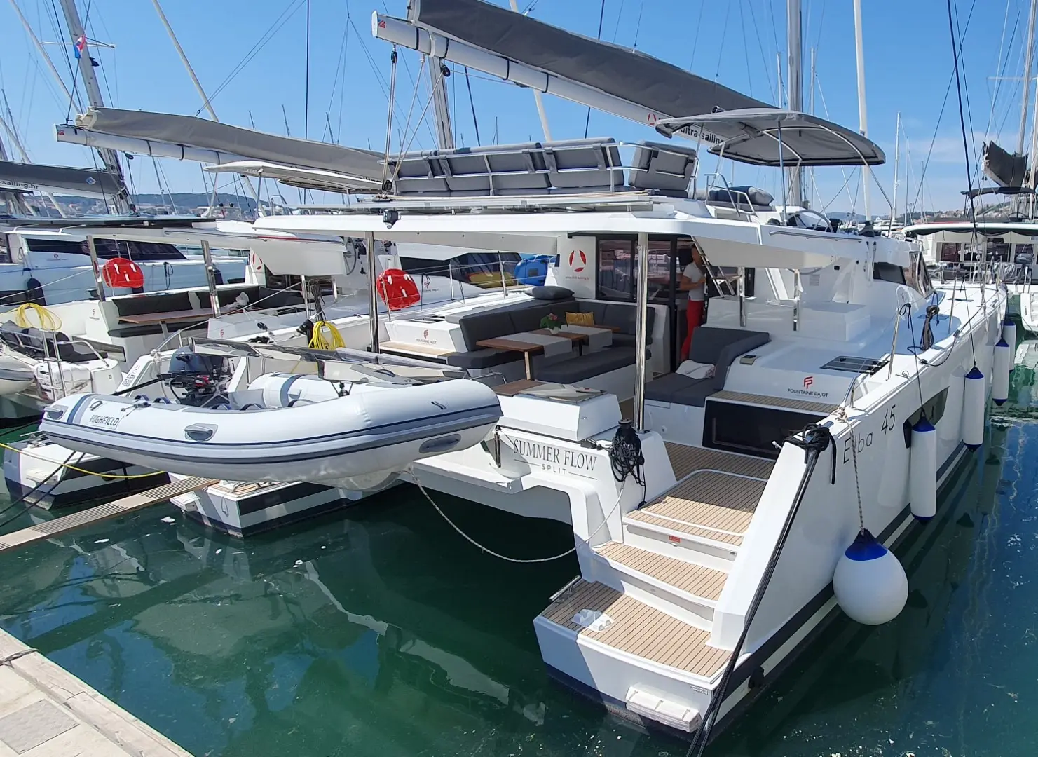 A white Fountaine Pajot Elba 45 catamaran named Endless Flow is docked in a marina, with a small inflatable boat at the back. The vessel has a shaded deck, outdoor seating, steps to the water, and is surrounded by other sailboats.