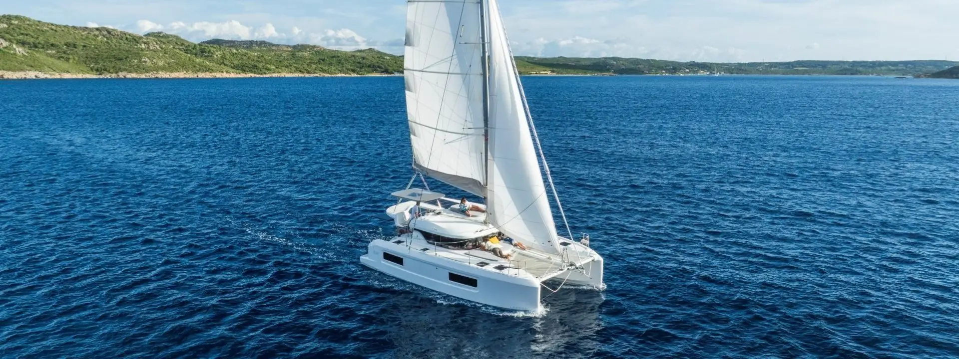 A white Lagoon 38 catamaran sailboat with raised sails glides across deep blue ocean waters near a green, hilly coastline under a partly cloudy sky.