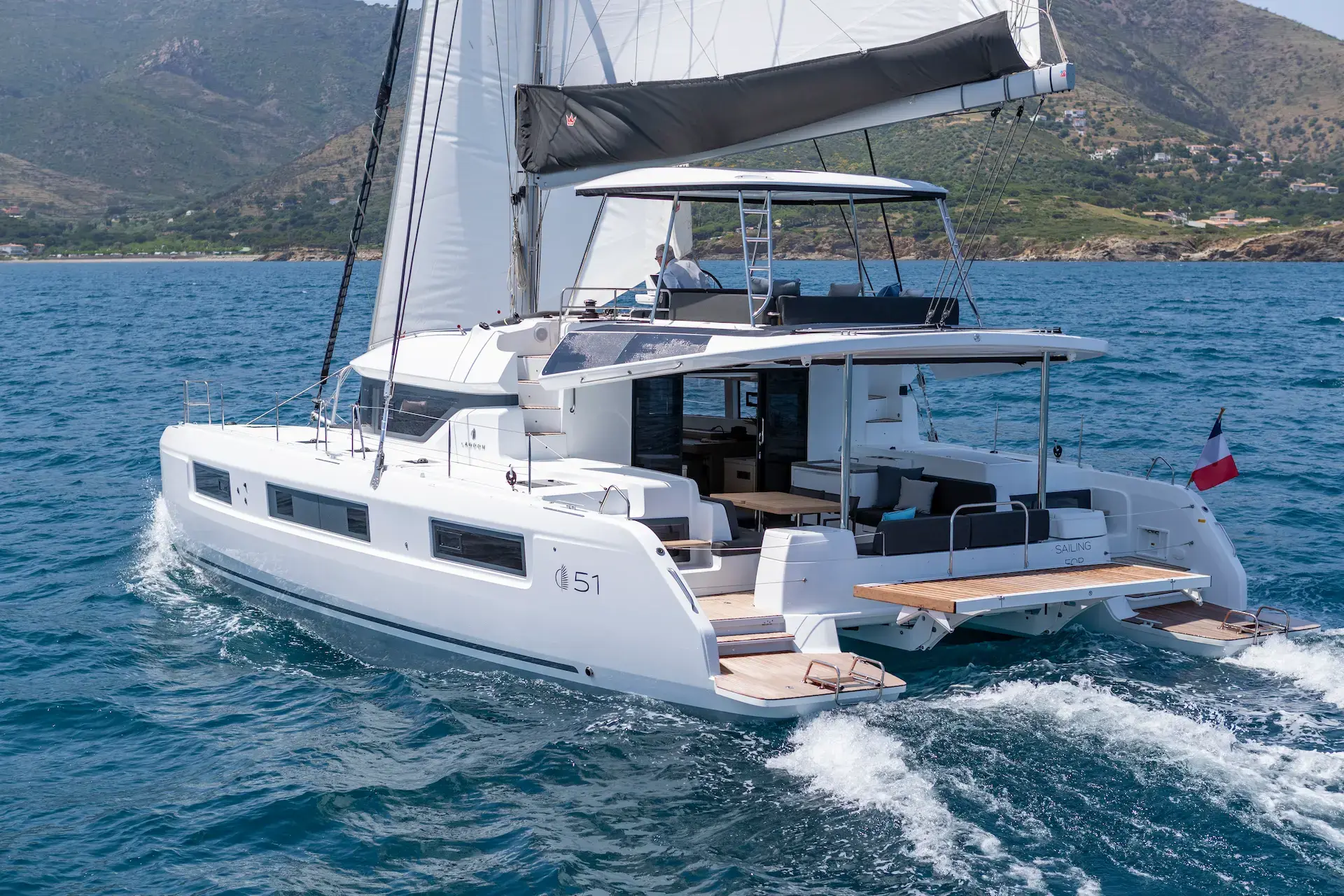 A modern white Lagoon 51 catamaran yacht with raised sails cruises on clear blue water near a mountainous coastline under a sunny sky.