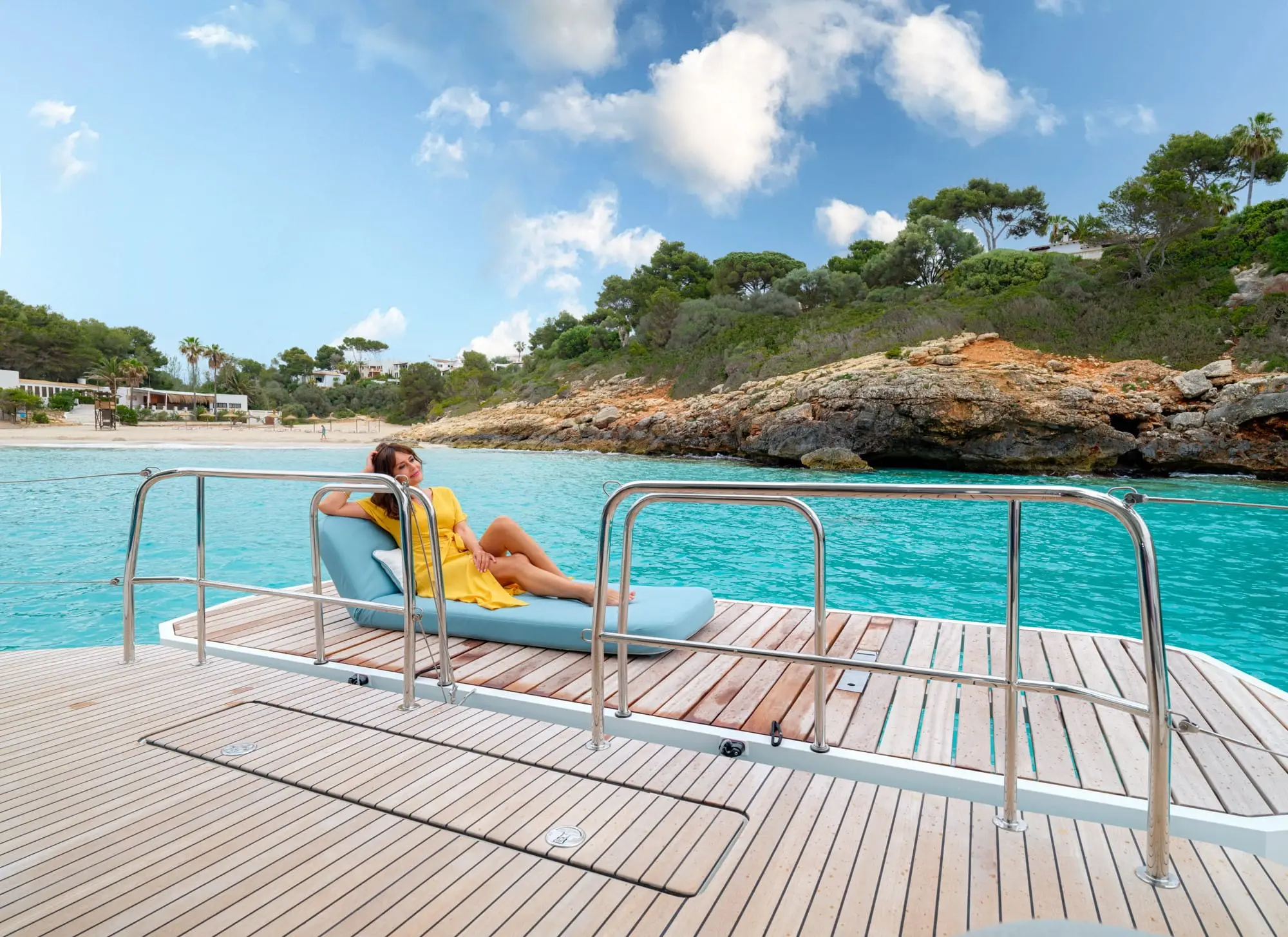 A woman in a yellow dress relaxes on a lounge chair on the wooden deck of a Lagoon 55 yacht, with turquoise water, rocky shore, trees, and a partly cloudy sky in the background.