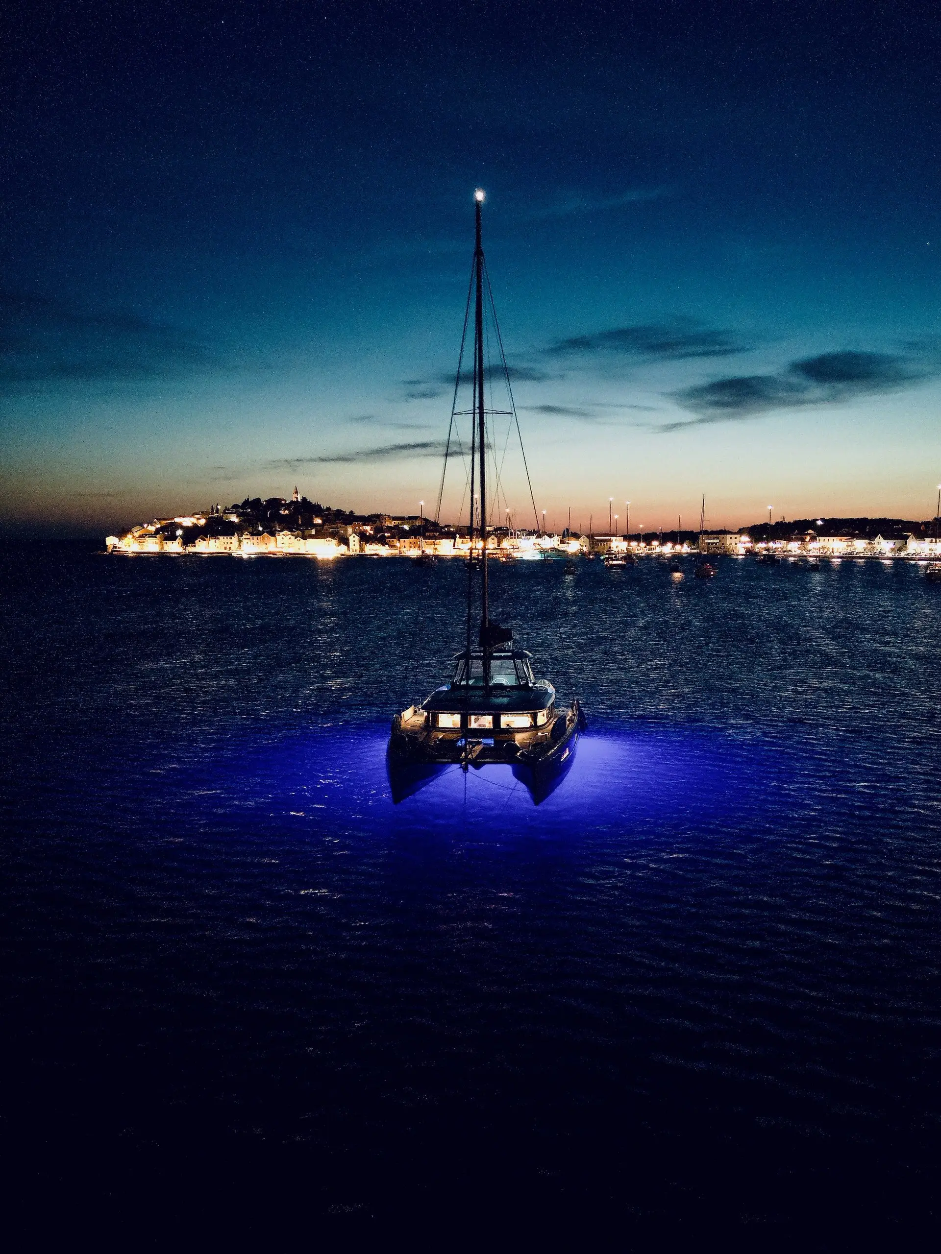 A Lagoon 46 sailboat with blue underwater lights floats on calm water at dusk, with a lit-up shoreline and distant boats visible under a darkening sky.
