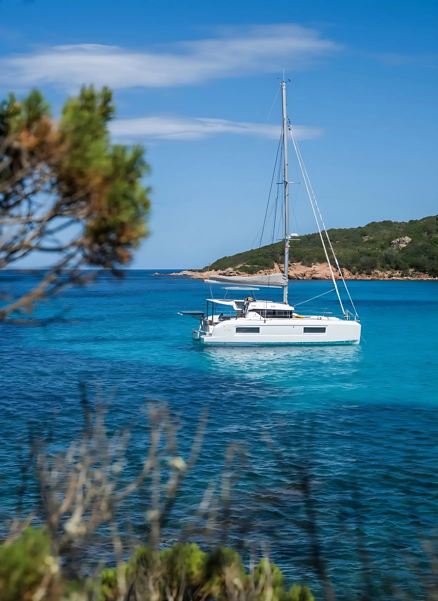 A white Lagoon 38 sailboat floats on clear turquoise water near a green, rocky coastline under a blue sky, partially framed by blurred branches and foliage in the foreground.
