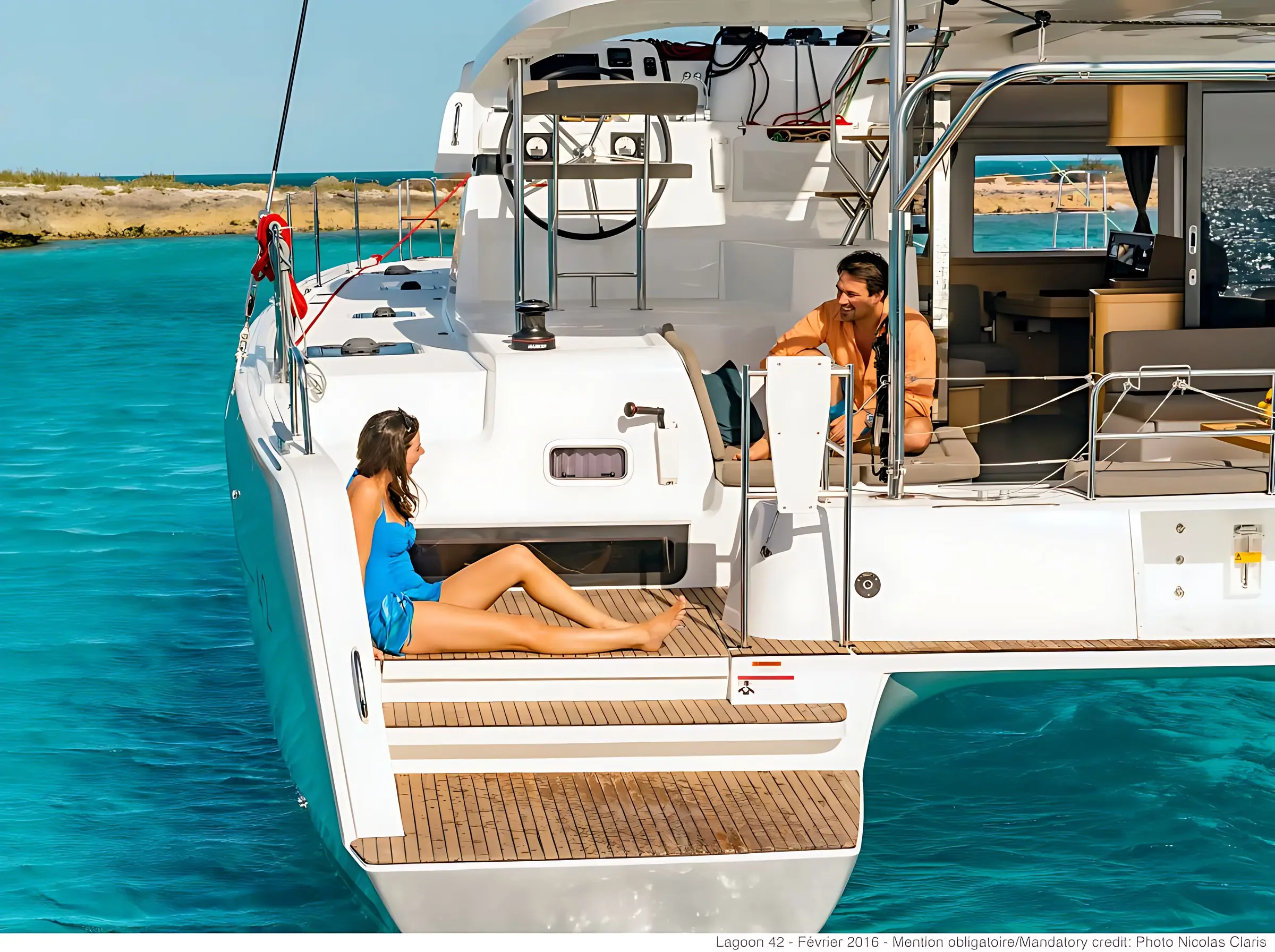 A woman in a blue swimsuit sits on the deck of a white yacht, facing a man in a tan shirt who is seated near the entrance. They are anchored in clear blue water near a rocky shoreline under sunny skies.