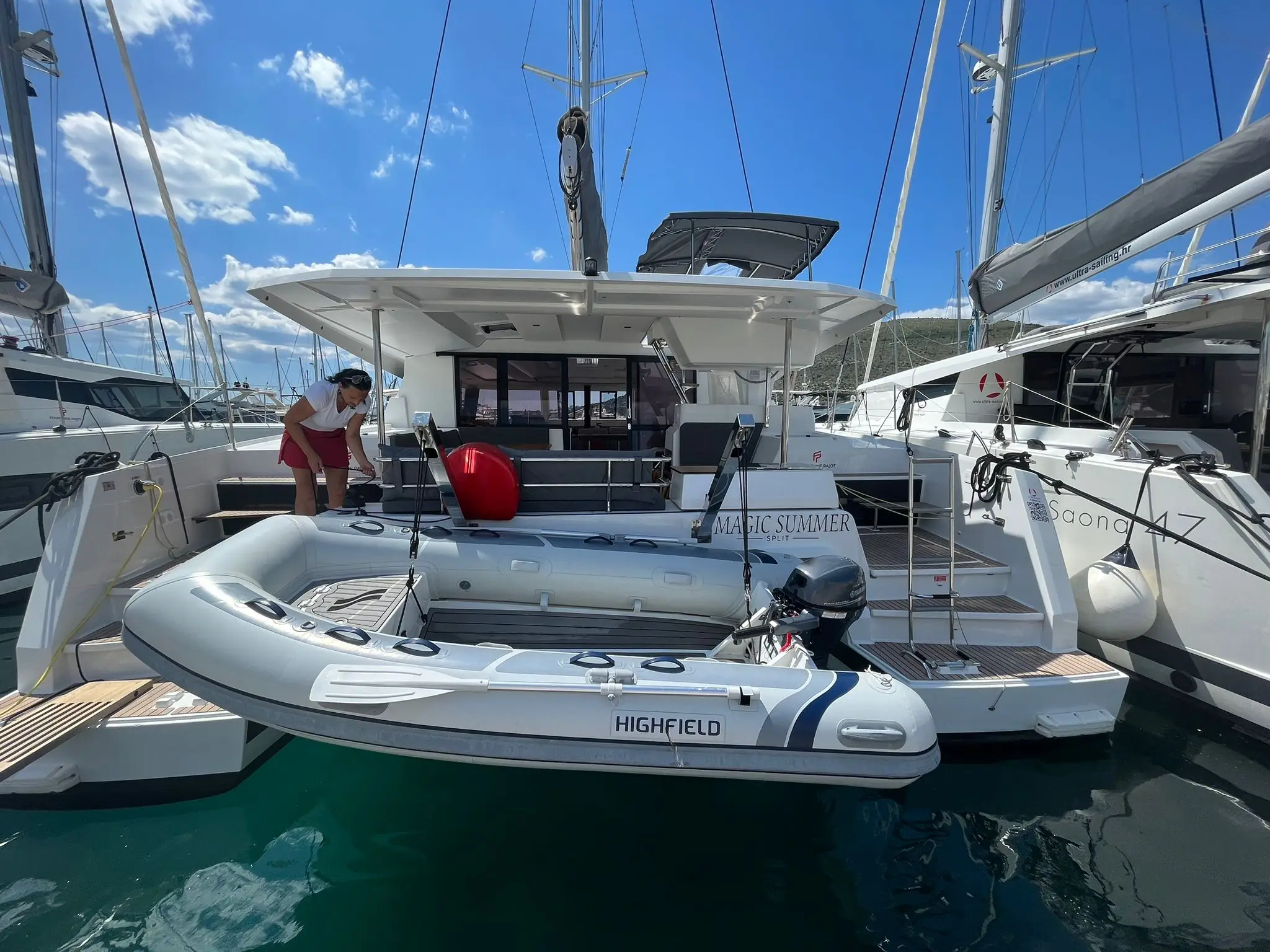 A woman stands on the deck of a Fountaine Pajot Astrea 42 yacht named Magic Summer, preparing a Highfield inflatable boat suspended over the water at a marina with other boats nearby under a clear blue sky.
