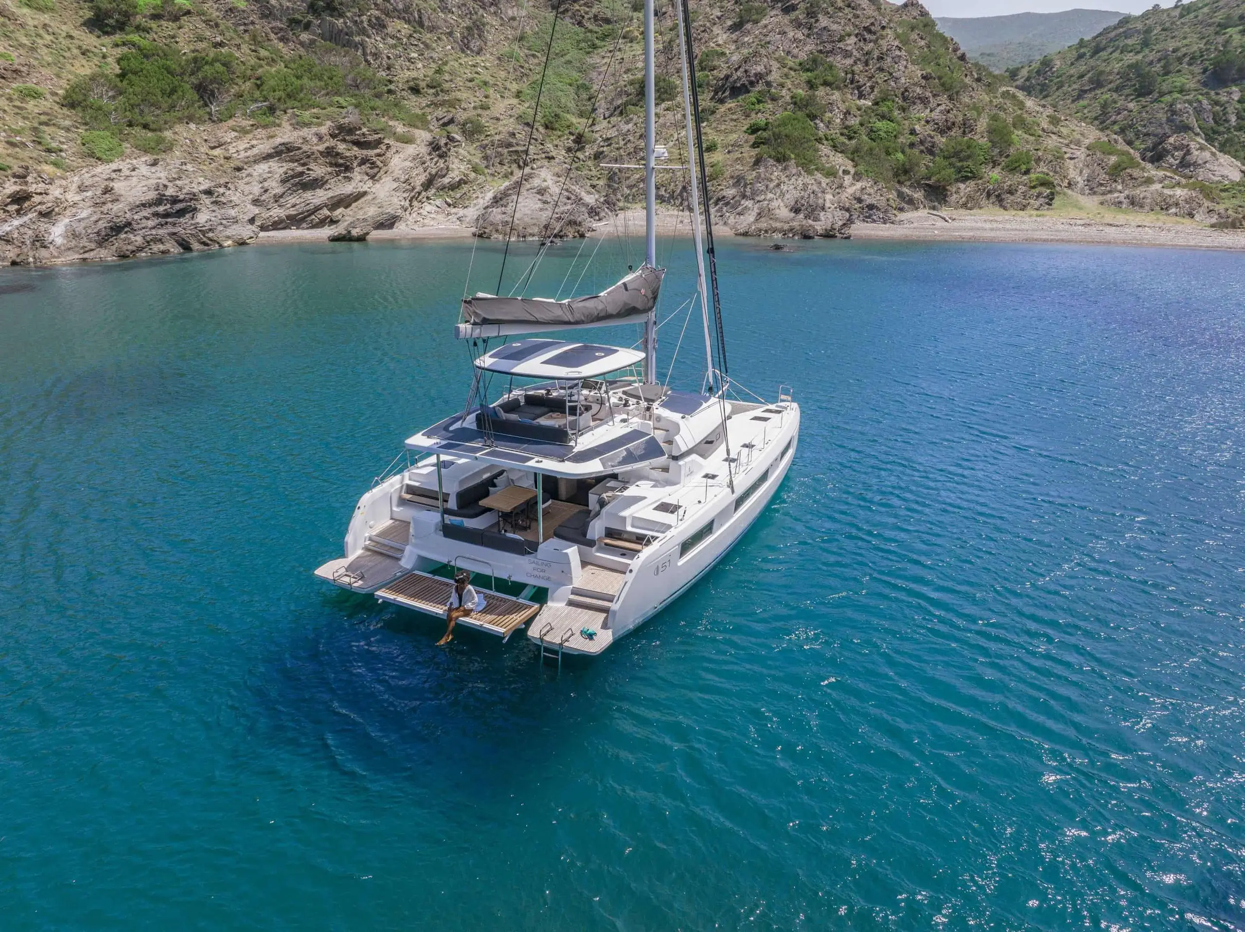 A white Lagoon 51 catamaran yacht is anchored in clear blue water near a rocky, green coastline, with a person sitting on the back platform enjoying the scenery.