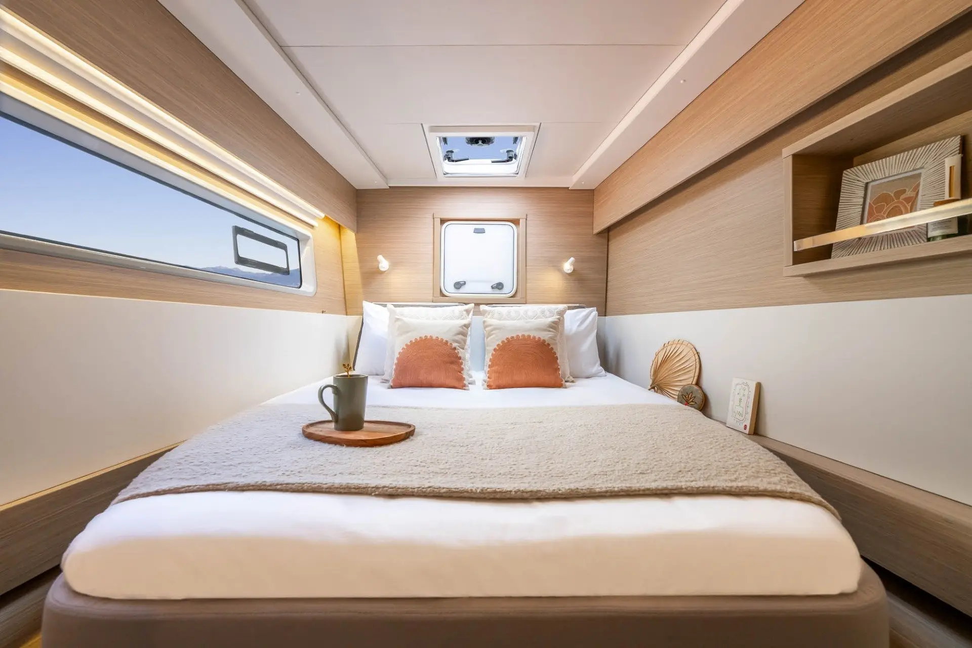 A cozy bedroom aboard the Lagoon 43 features a neatly made double bed, beige and white decor, two pillows, a tray with a mug, a window above the bed, a side shelf with books, and soft natural light streaming in.