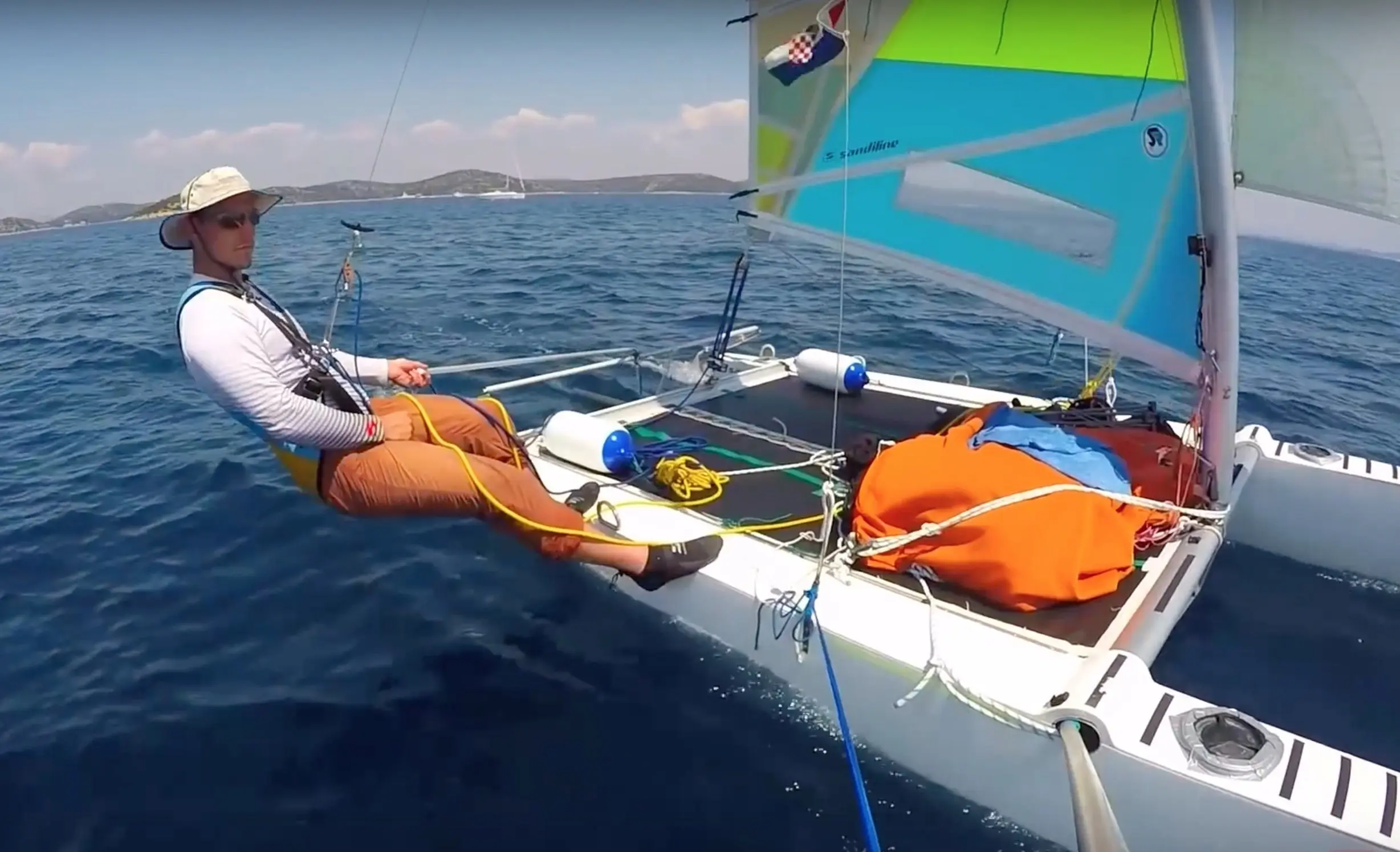 Captain Igor Stropnik sailing a small catamaran with a colourful sail on open blue sea, islands on the horizon.