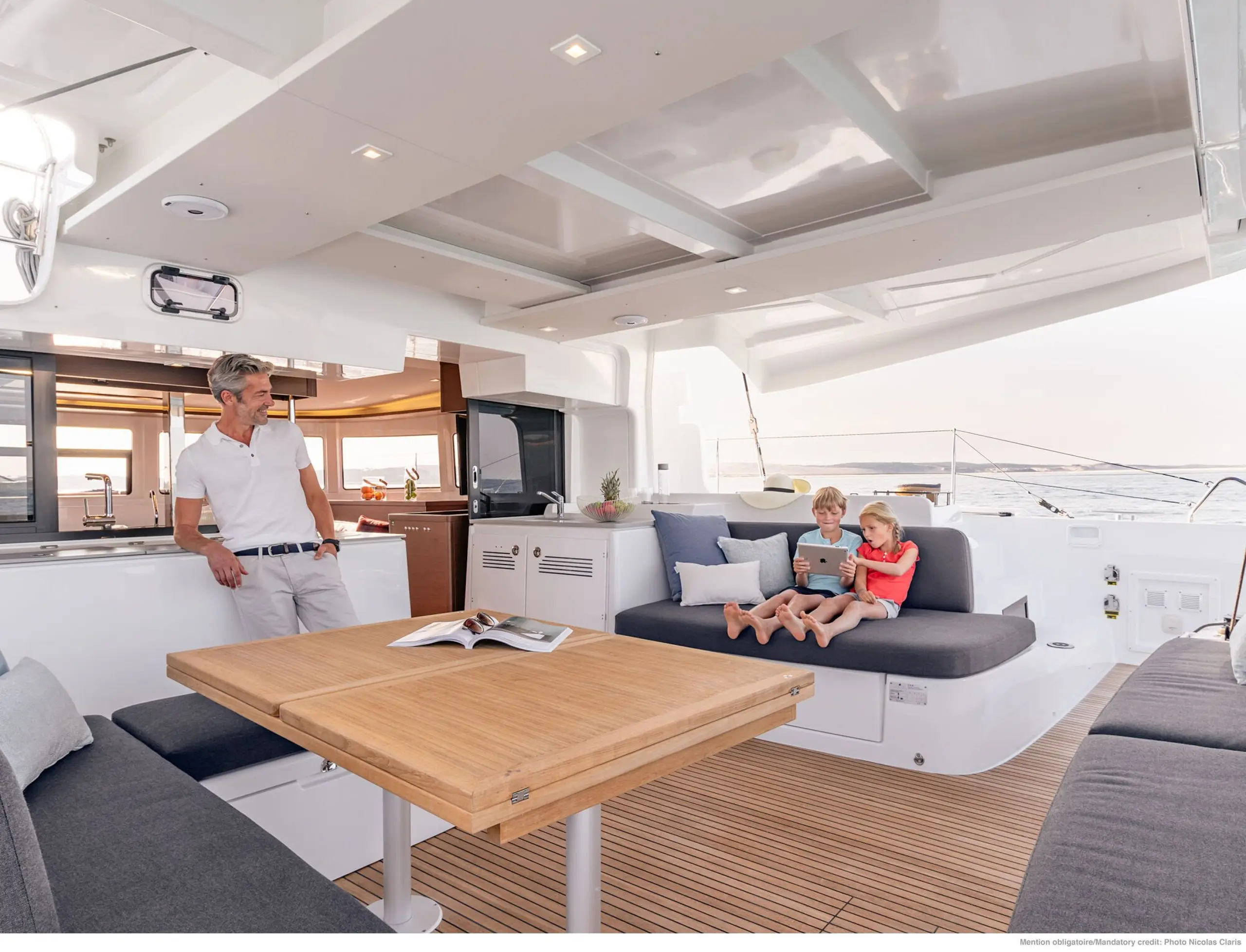 A man stands near a wooden table on a Lagoon 46 yacht while two children sit on a cushioned bench, one using a tablet. The setting is bright and modern, with the sea visible in the background.
