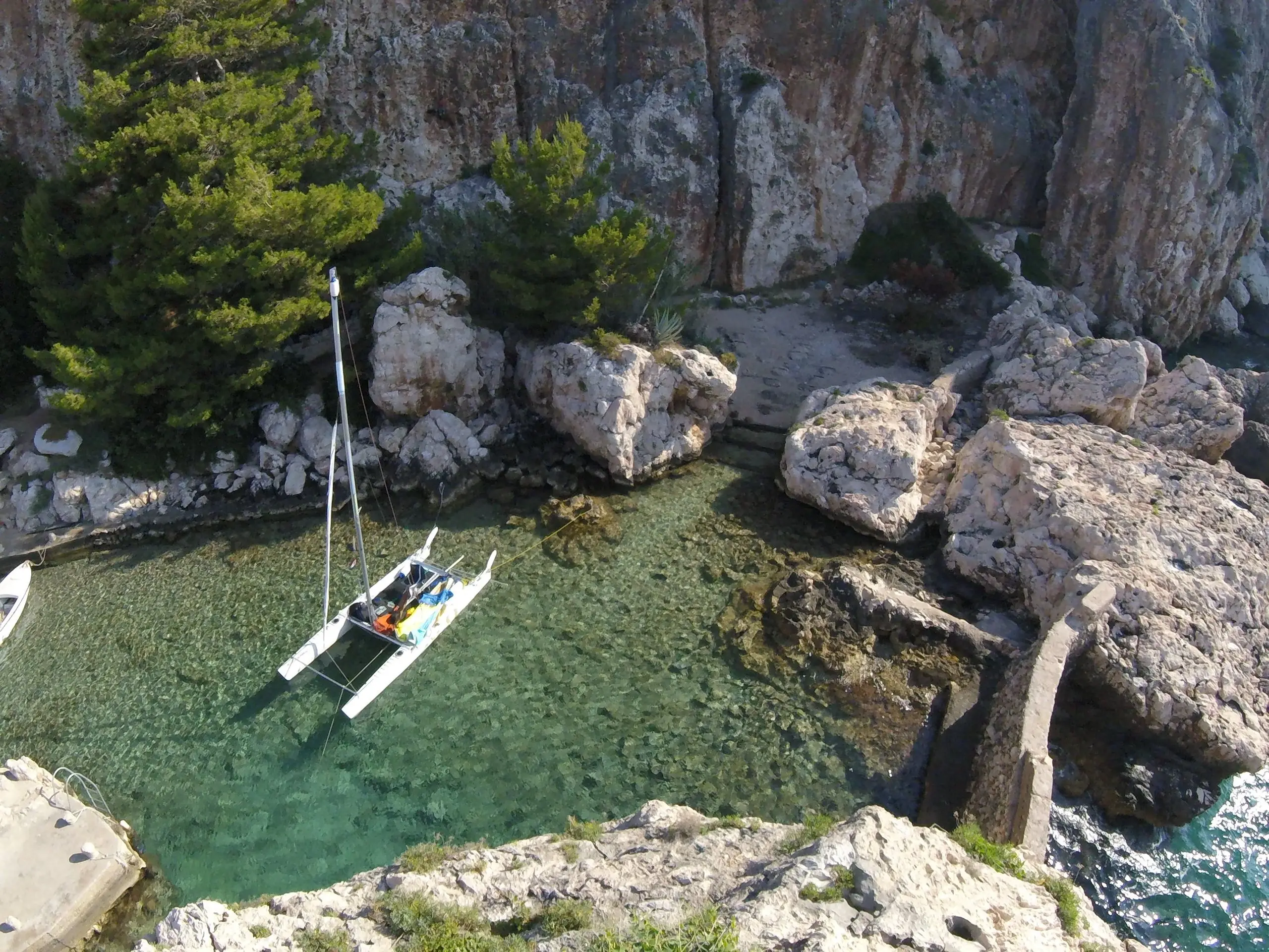 A small sailing catamaran moored in a clear turquoise cove below white limestone cliffs and pine trees.