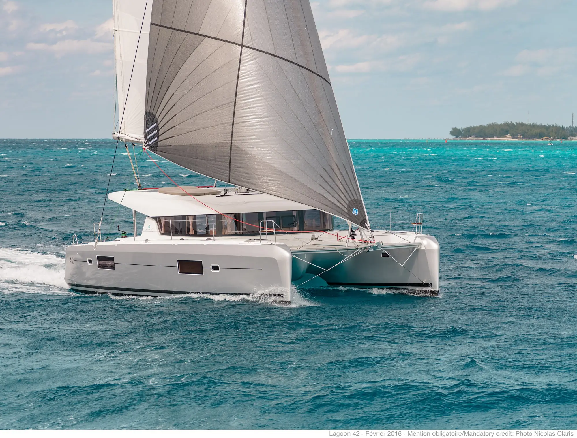 A modern white catamaran sails quickly across turquoise ocean water under a large gray and white sail, with a distant shoreline visible on the horizon.
