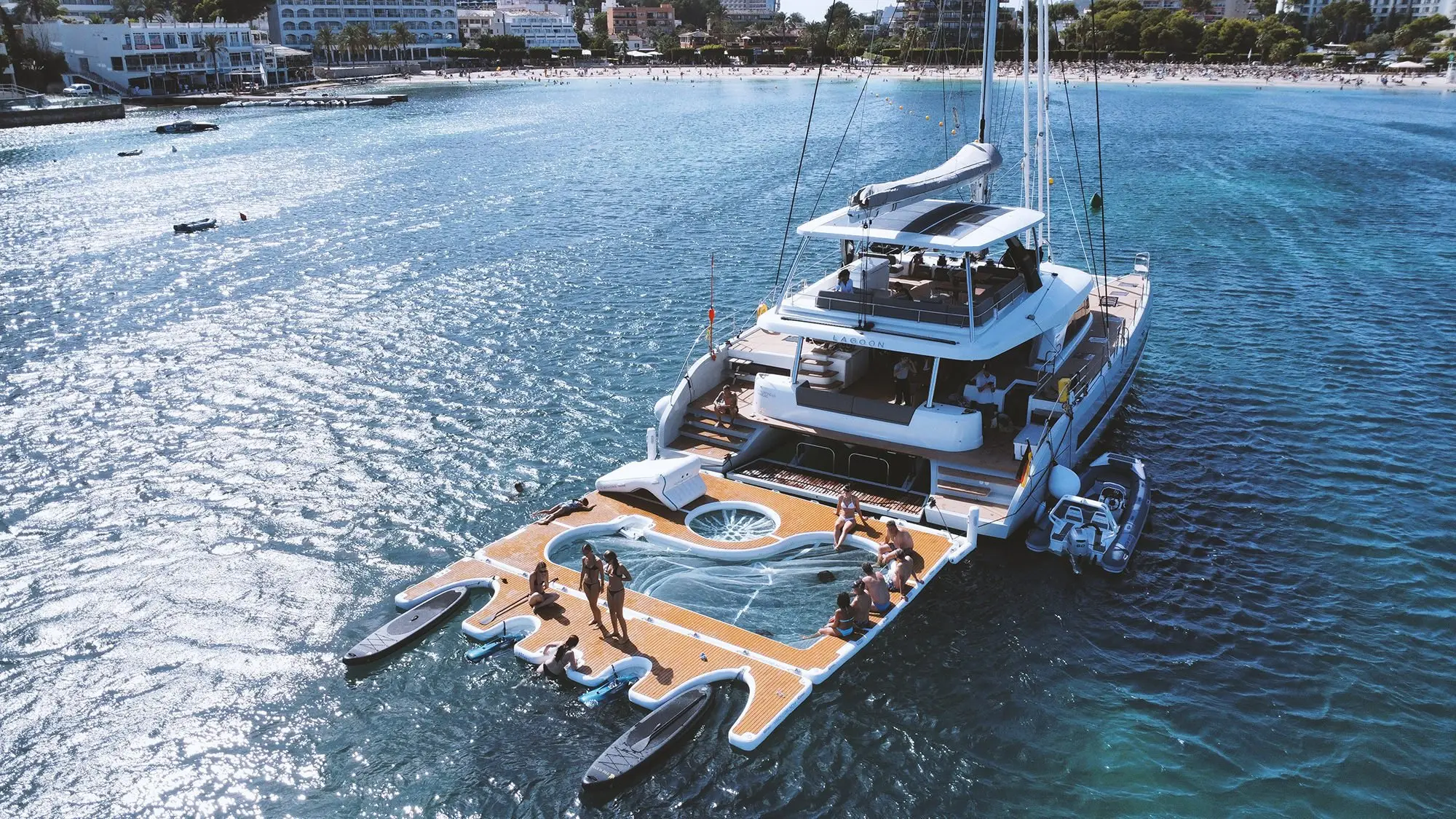 A large, luxurious Lagoon Sixty 5 catamaran yacht is anchored near the shore, with people relaxing on its spacious deck and floating platform. Paddleboards and an inflatable lounge are set up beside the boat on the clear blue water.