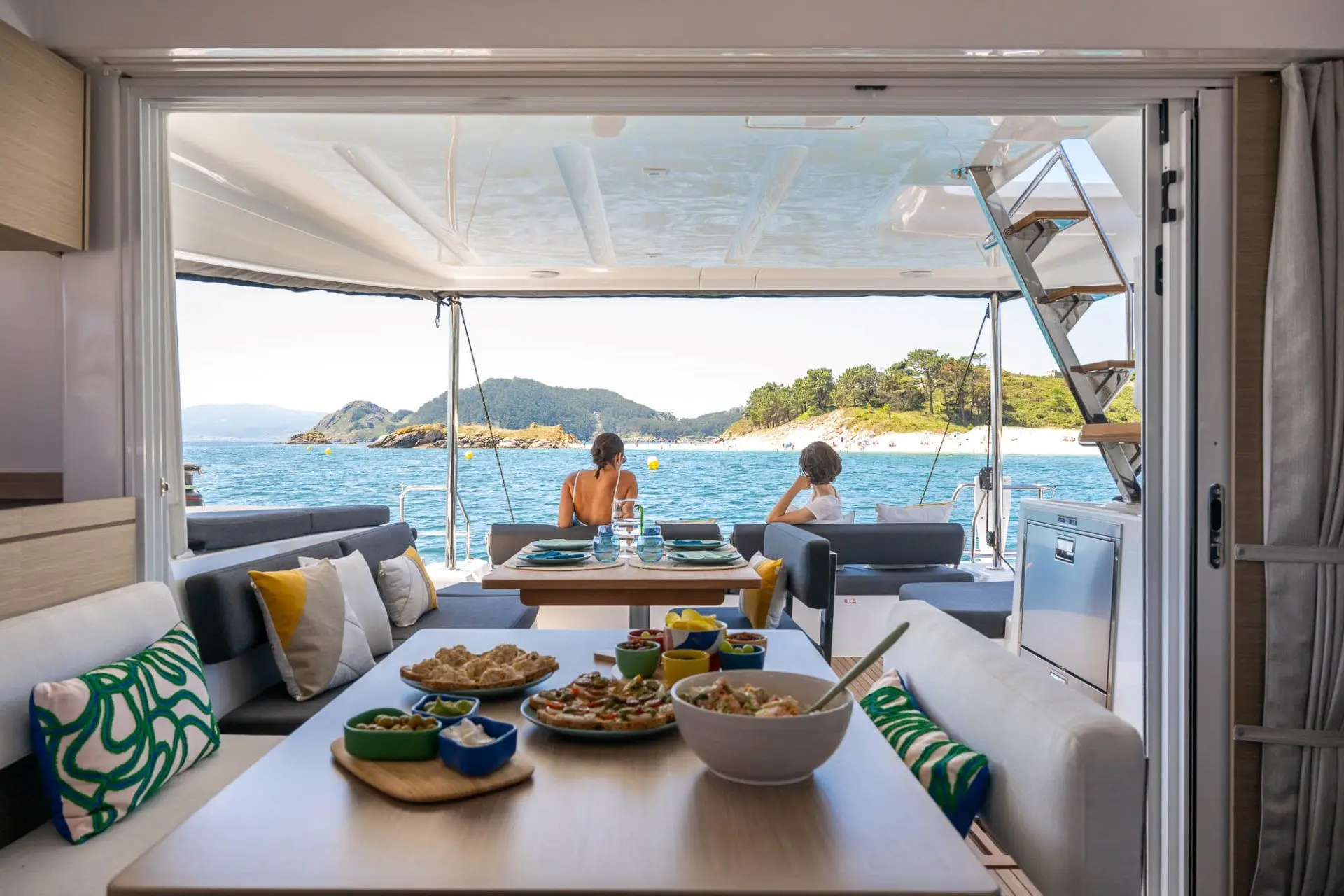 A table set with various dishes is inside a Lagoon 43 boat cabin, while two people sit at the back enjoying the ocean view and nearby islands under a sunny sky.