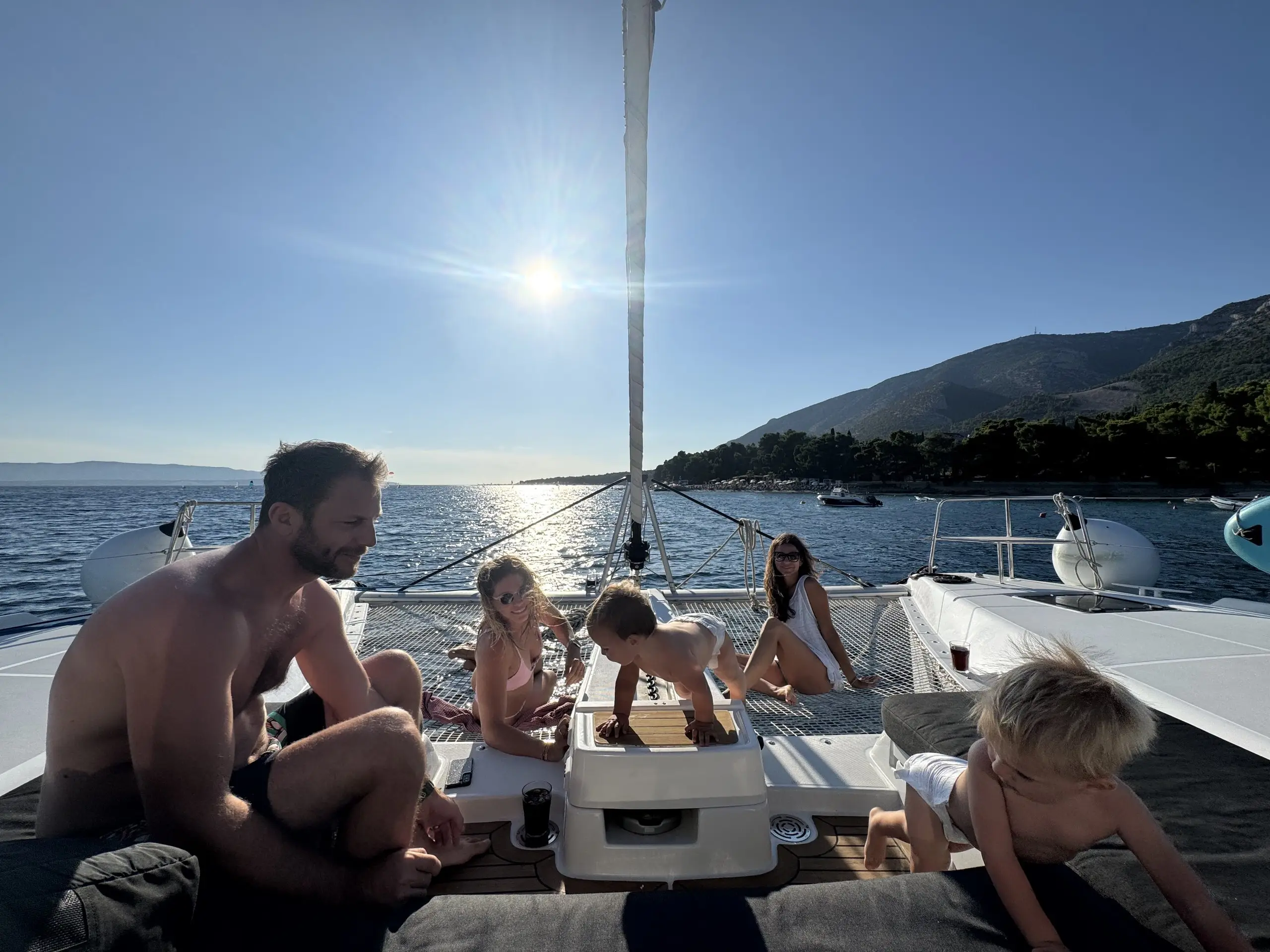 A family relaxes on the deck of a Lagoon 46 sailboat in sunny weather, with two children playing and two adults watching. The boat glides over calm water, framed by mountains and a clear blue sky in the background.
