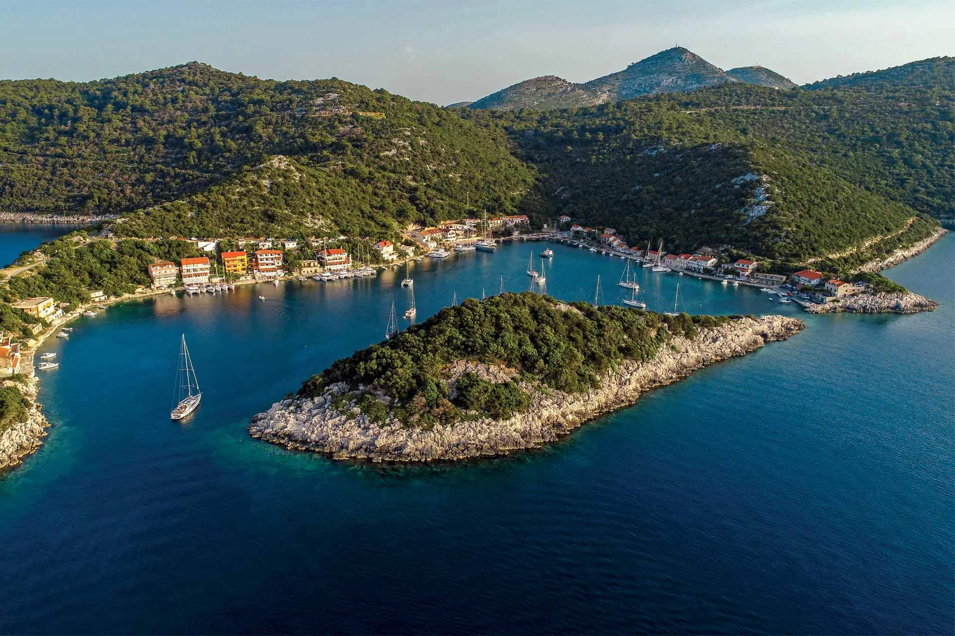 Aerial view of a small, lush green island surrounded by blue water, with Lagoon 51 sailboats anchored nearby and a coastal village nestled among hills in the background.