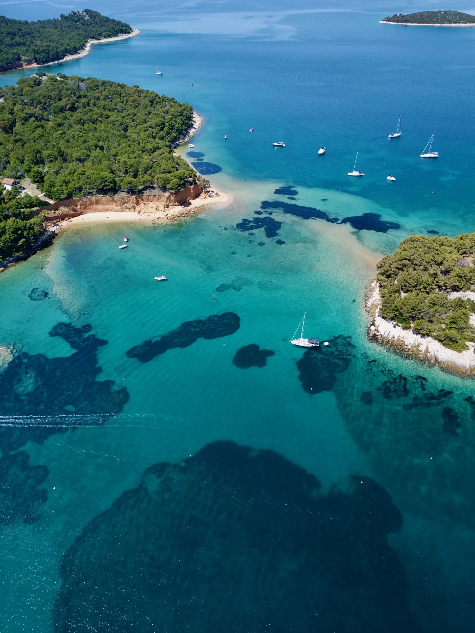 Aerial view of a turquoise bay with clear water, sandy beaches, lush green forests, and several Lagoon 46 sailboats anchored near the shore.