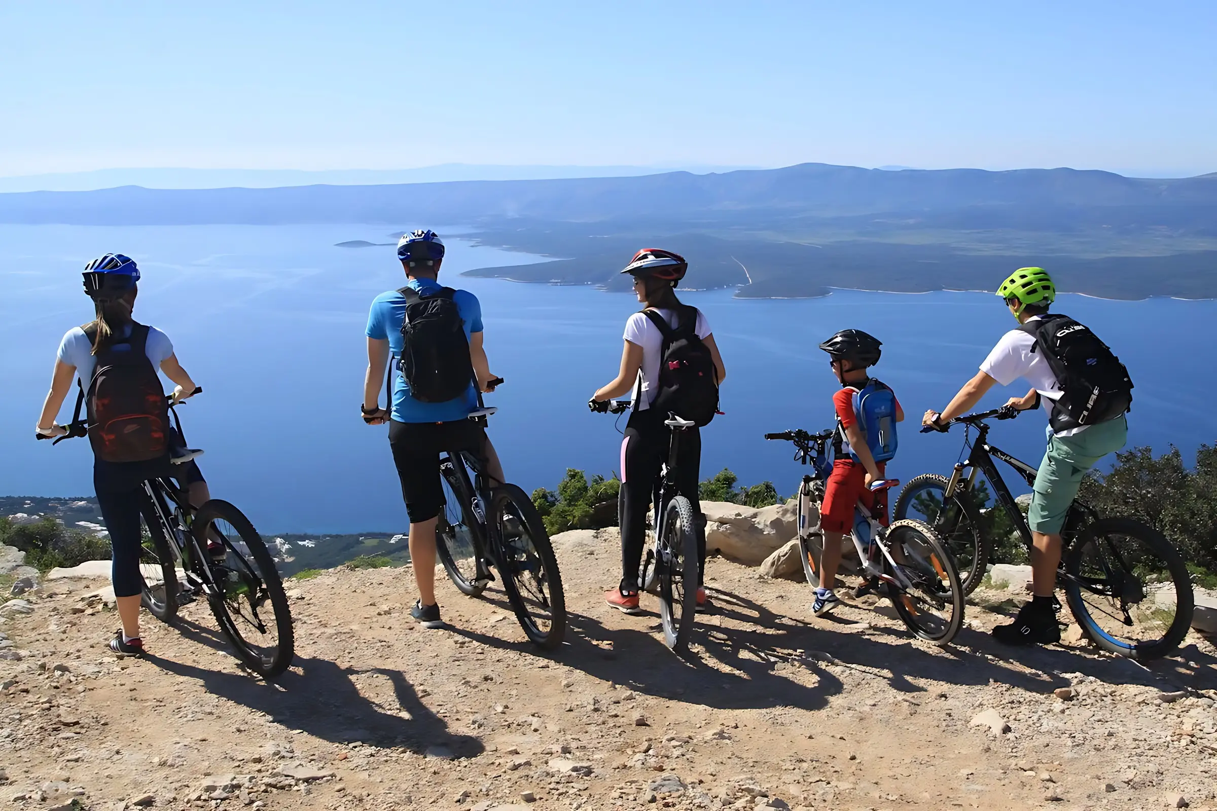Five cyclists wearing helmets and backpacks pause at a scenic viewpoint, overlooking a vast blue body of water and distant mountains under a clear sky.