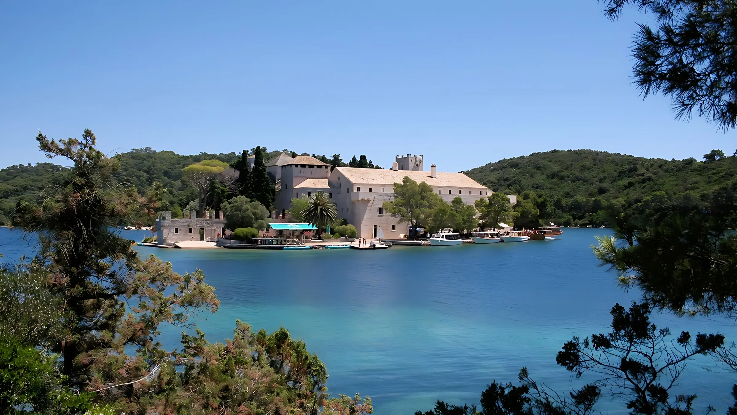 A large stone monastery with a tower sits on a lush, tree-covered island surrounded by clear blue water under a sunny, cloudless sky. Boats are docked along the shoreline.