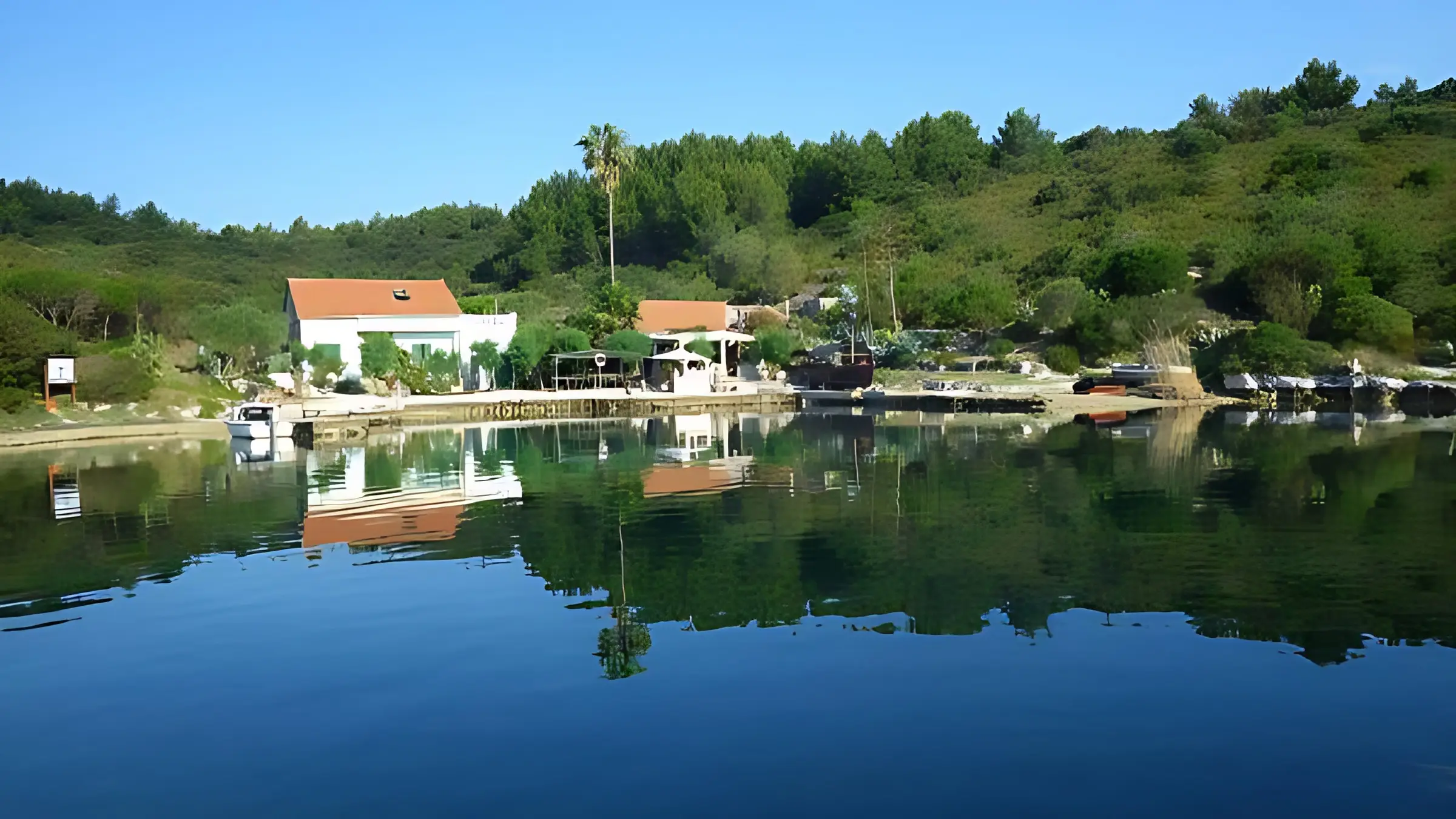 A white house with a red roof sits near the edge of a calm, reflective body of water—perfect for exploring some of the best catamaran routes—surrounded by green trees and shrubs under a clear blue sky. A small dock and boats are visible by the shore.