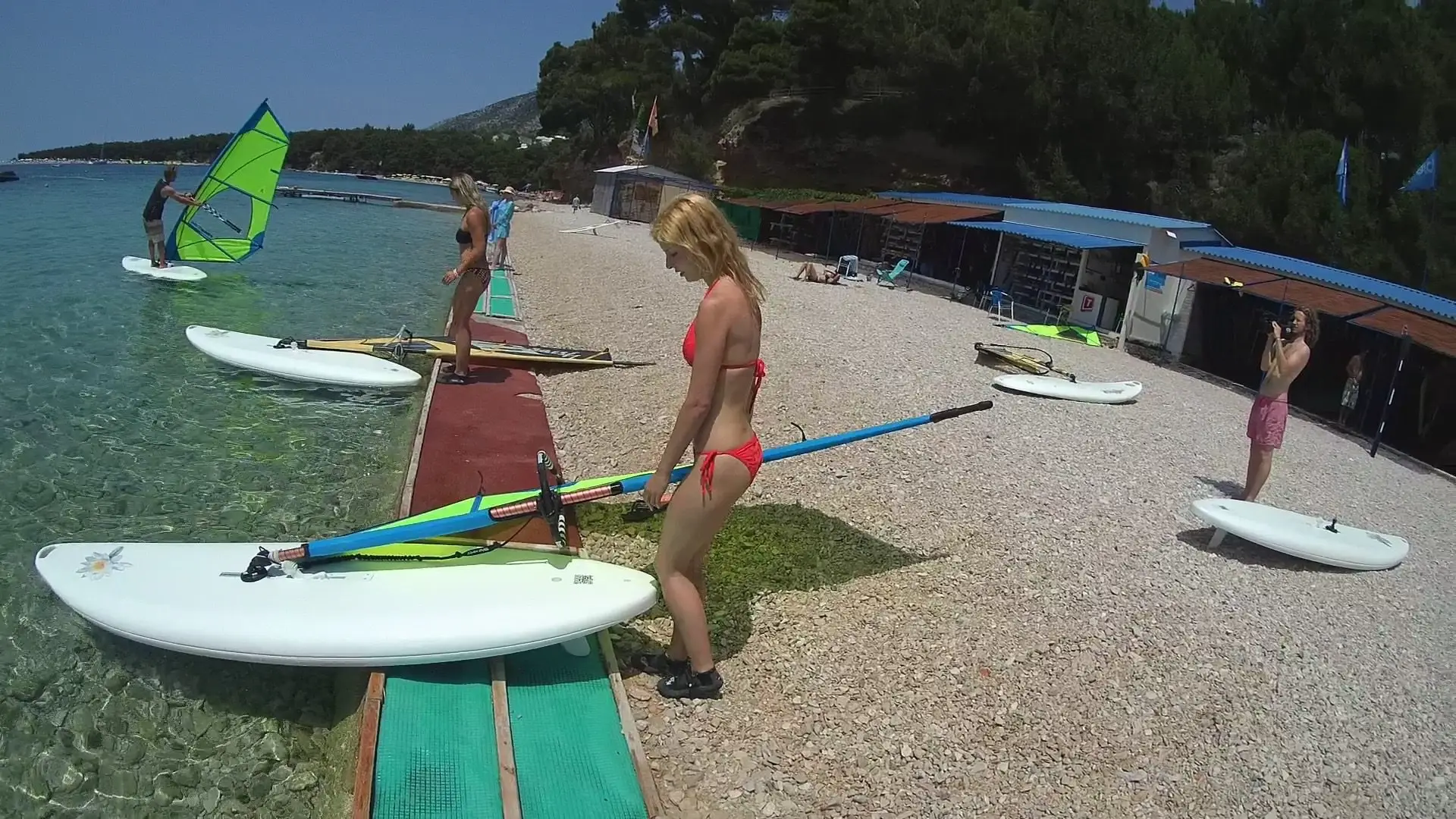 Several people in swimwear prepare windsurfing boards on a pebbled beach by clear blue water. Trees and beach facilities are in the background under a sunny sky.