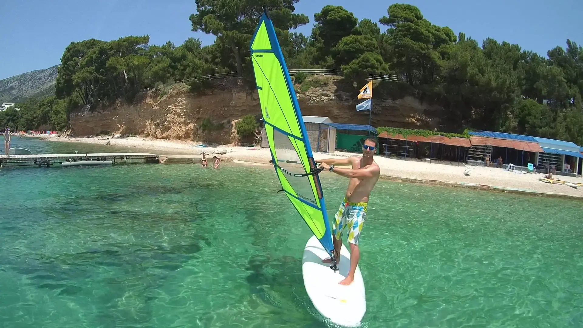 A man in colorful swim trunks windsurfing on clear turquoise water near a sandy beach with trees and small buildings in the background.