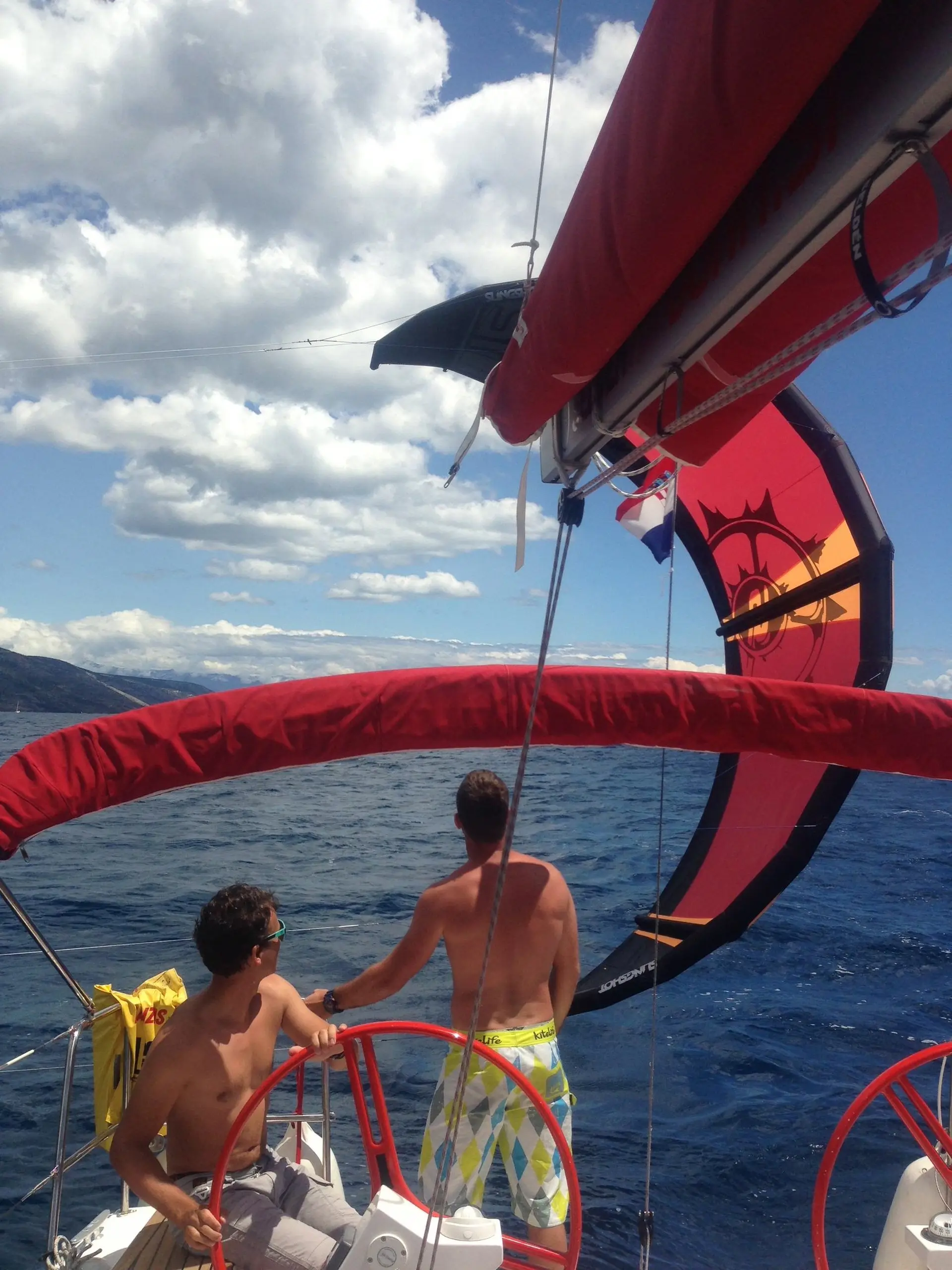 Two people on a sailboat look out at the sea under a partly cloudy sky. A colorful kite or kite sail is visible in the background above the water. The scene suggests a sunny, leisurely day on the ocean.
