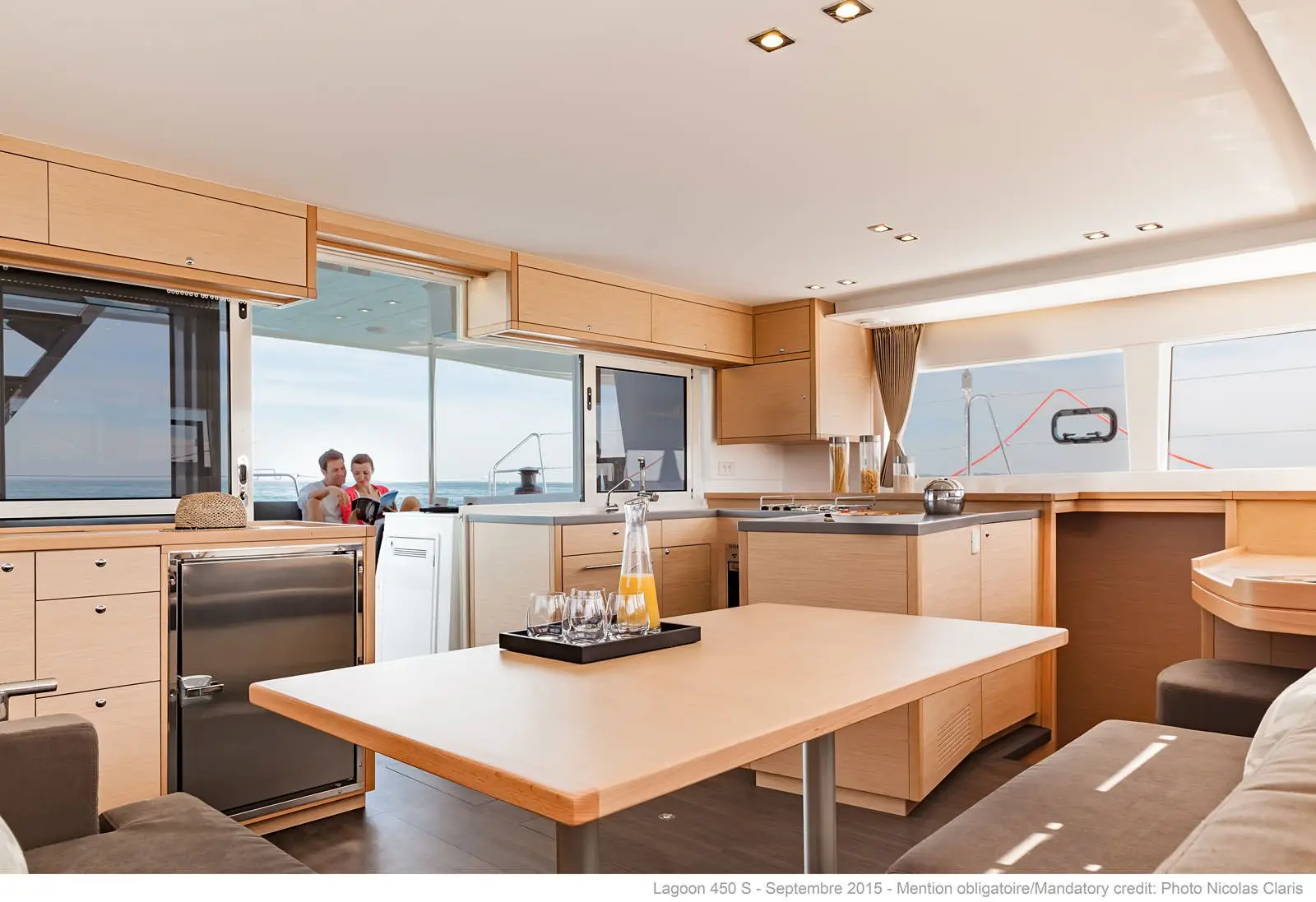 The interior of a modern yacht with light wood cabinets, a dining table, kitchen area, and large windows. Two people are visible outside on the deck, with the ocean in the background.