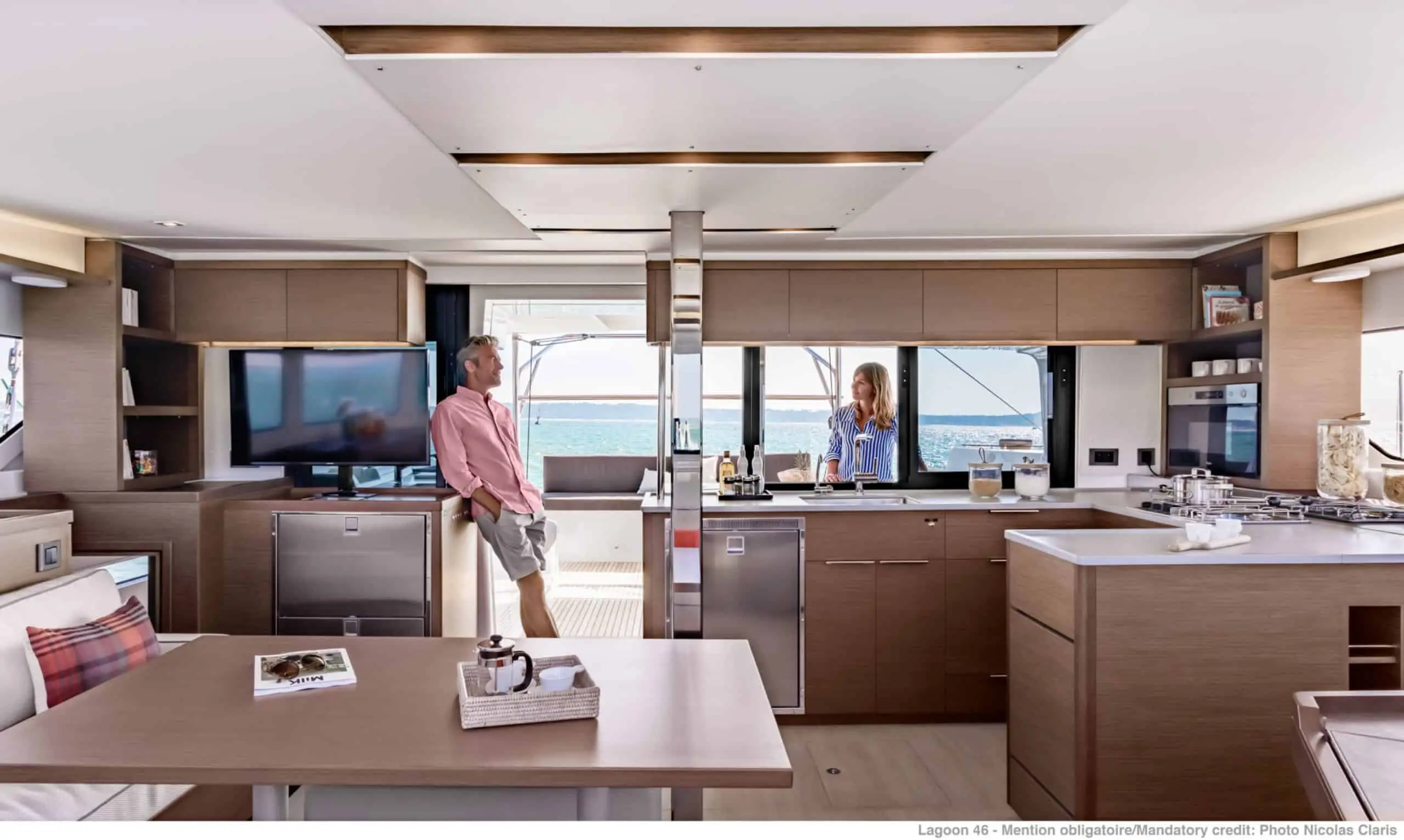 A man and woman converse inside a modern Lagoon 46 yacht kitchen and lounge area, featuring light wood cabinetry, large windows with a view of the sea, and stylish appliances.