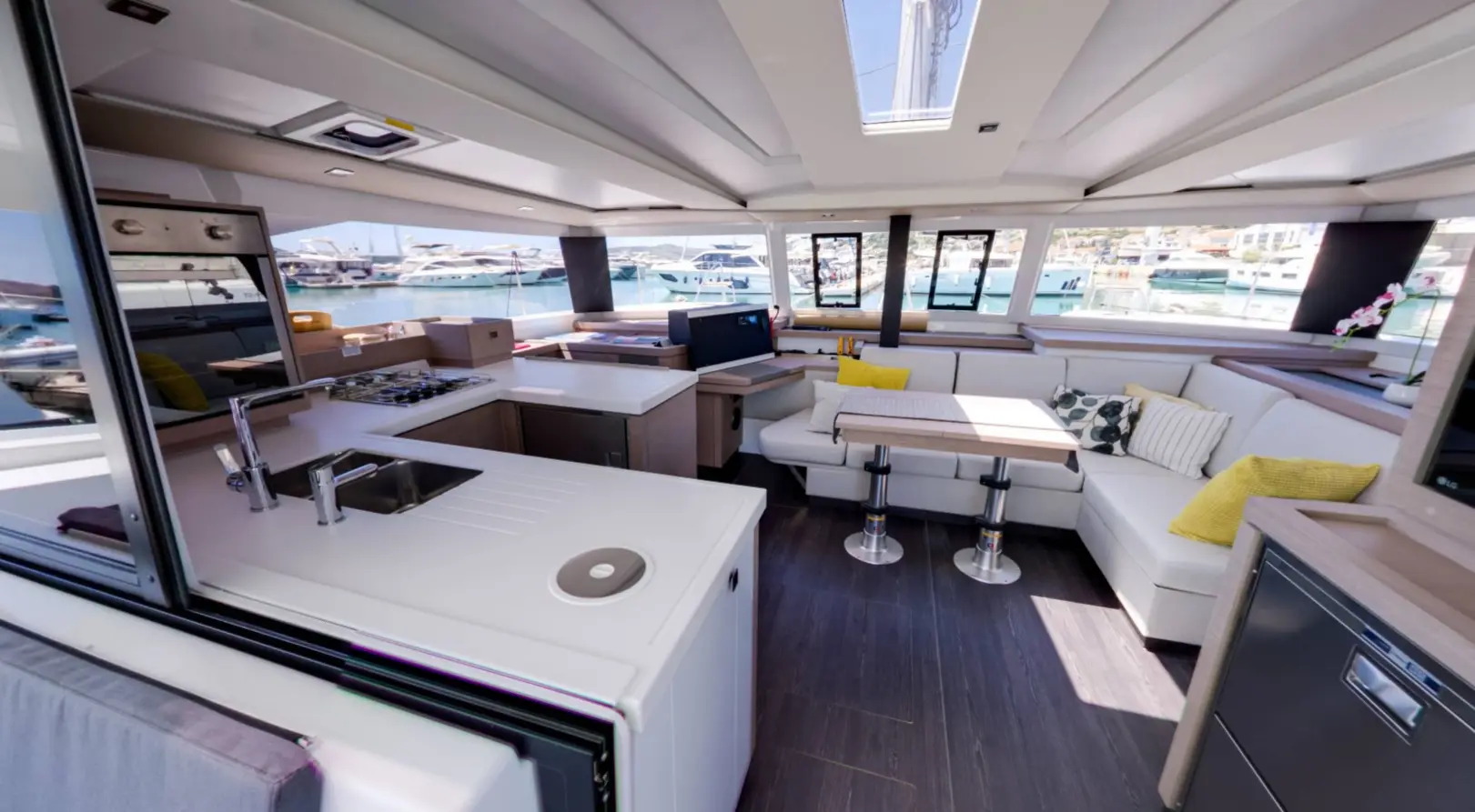The interior of this modern Fountaine Pajot Astrea 42 yacht features a sleek kitchen area, stove, seating, dining tables, TV, and large windows overlooking the marina. The space is bright and contemporary with neutral decor.